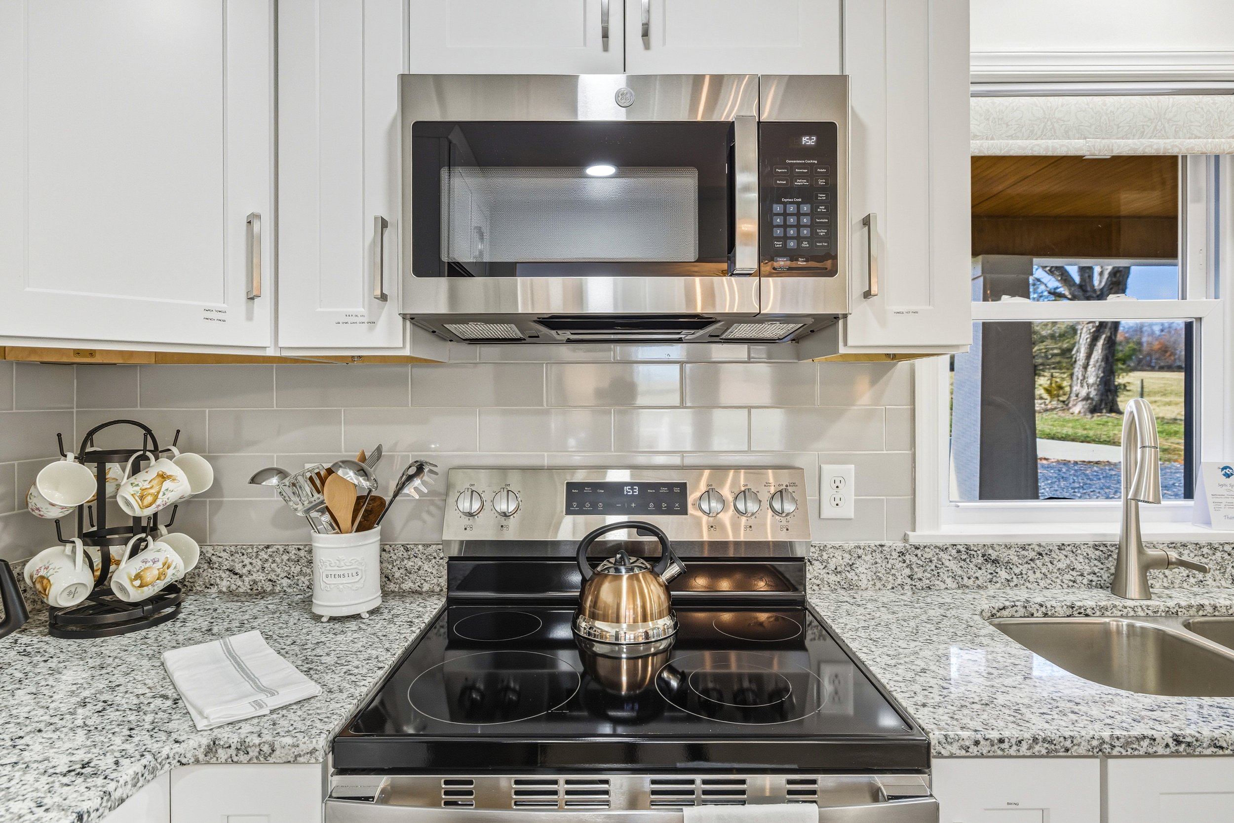 Kitchen with white cabinetry, granite countertops, and stainless steel appliances including a microwave above a stove with a kettle on the cooktop. There are white cups with a floral design on a black metal stand, utensils in a white holder, and a di