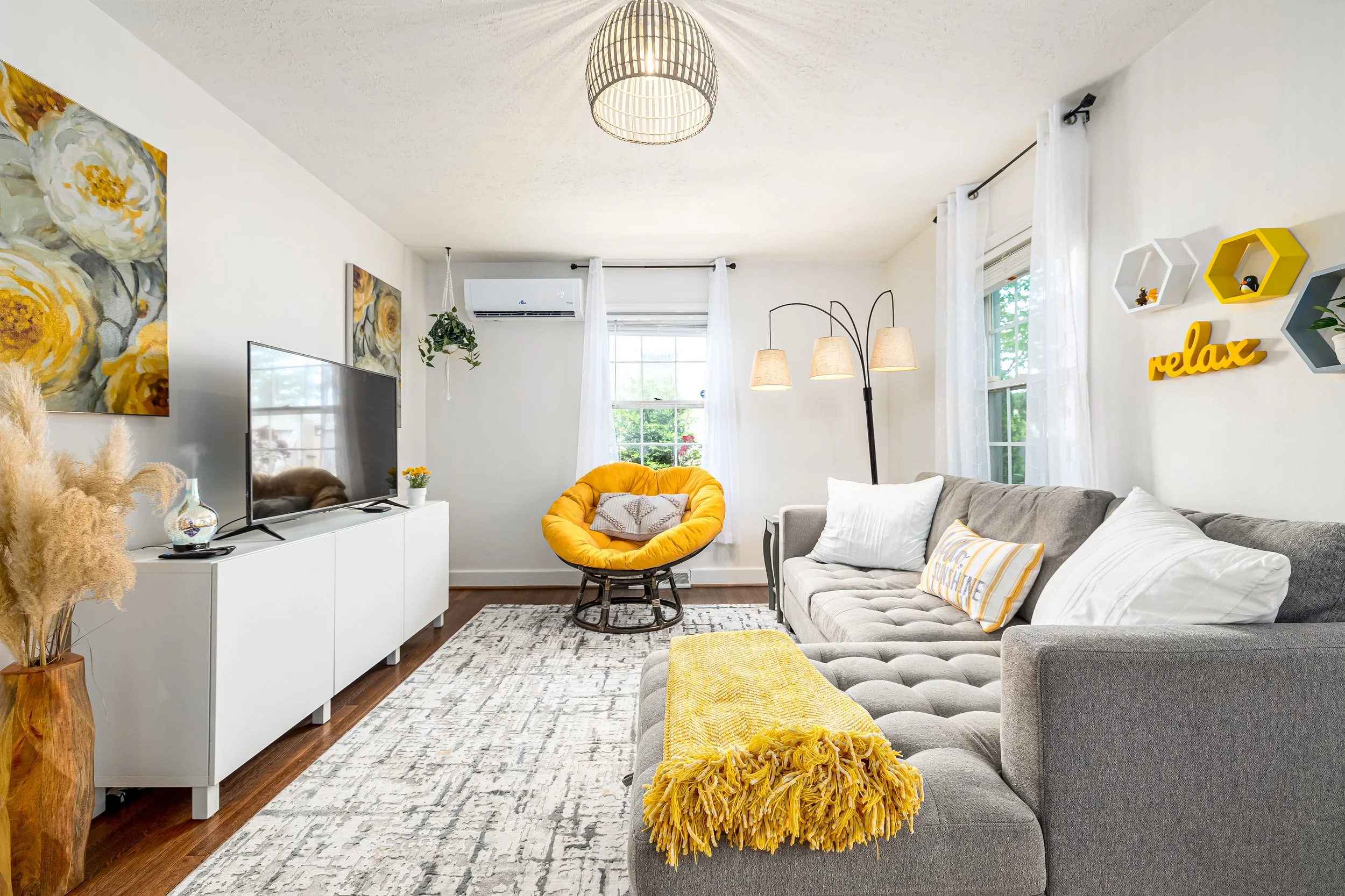 Living room with a gray sofa, yellow throw pillow and blanket, yellow circular chair, white TV stand with a flat-screen TV, wall art with yellow flowers, white walls, windows with white curtains, a white carpet rug, hanging light fixtures, decorative