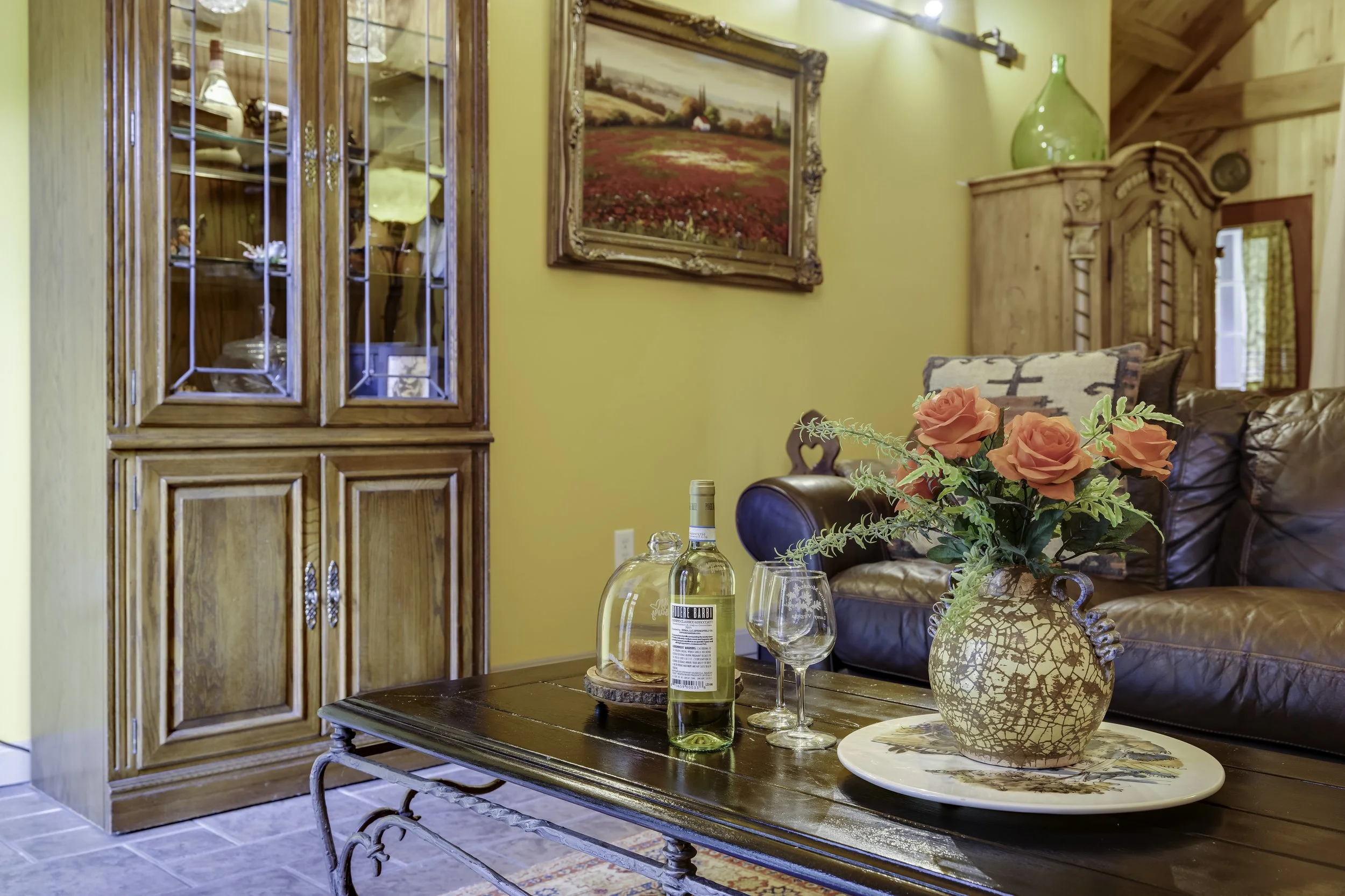 A cozy living room with a wooden table, glass bottles of wine, a large beige vase with orange roses, and a brown leather sofa. There is a large framed landscape painting on a yellow wall and a wooden cabinet with glass doors displaying decorative items.