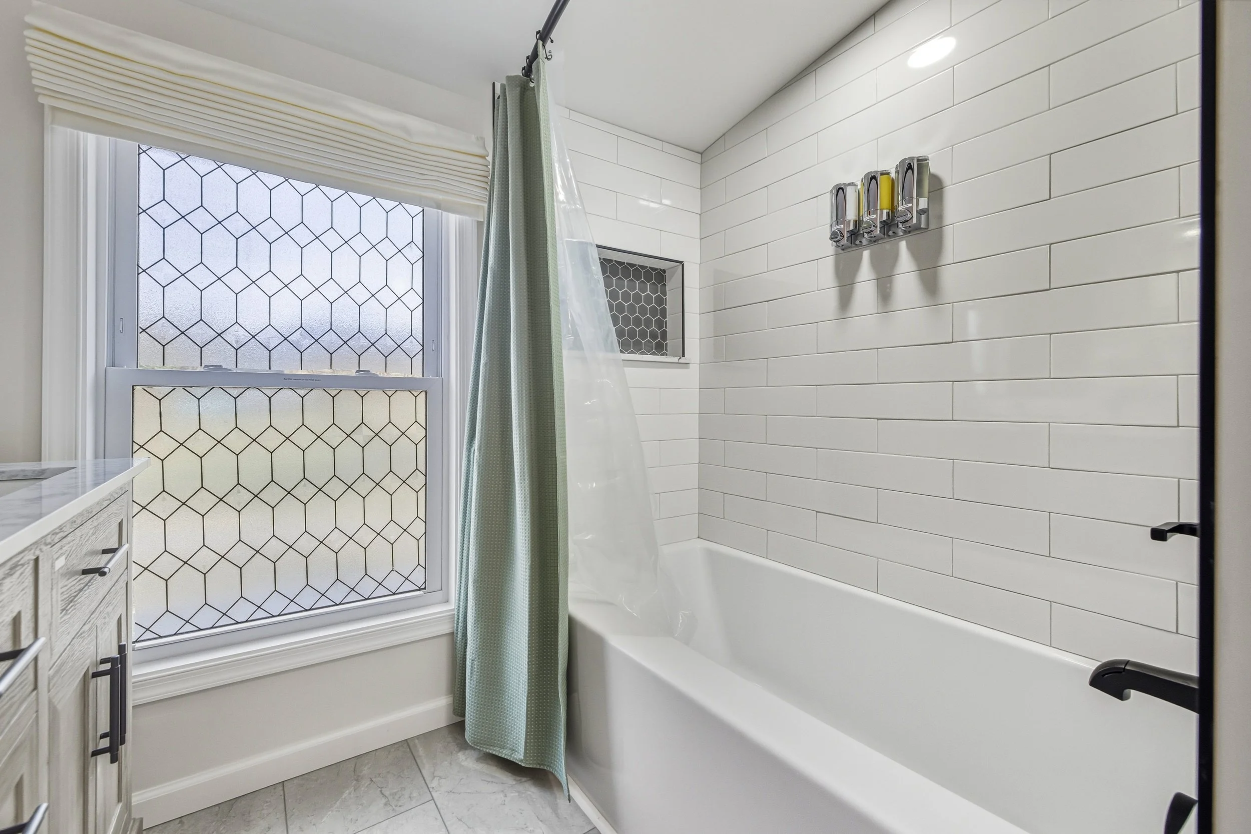 A bathroom with a white bathtub, white subway tiles on the wall, a window with geometric patterned frosted glass, a shower curtain, and a small built-in shower niche.