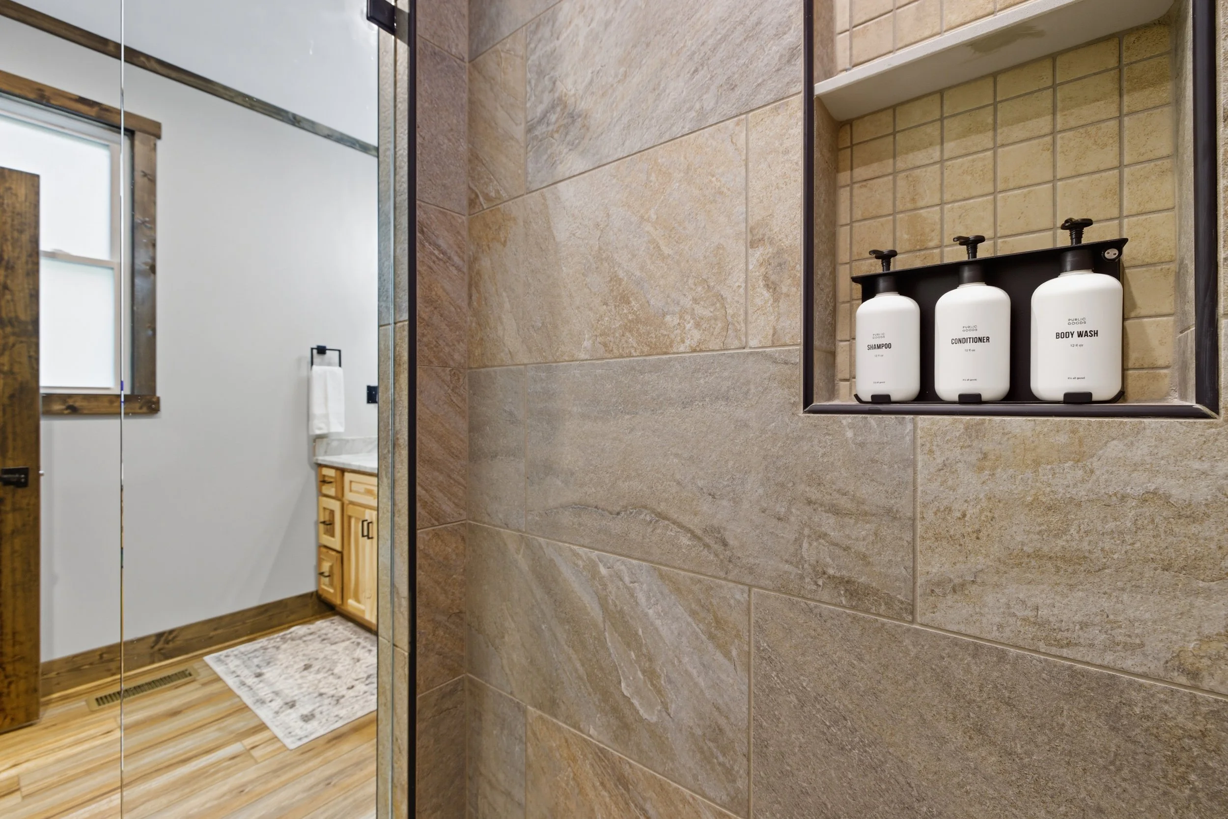A bathroom shower area with beige and brown stone tiles. Inside the shower, there is a built-in niche with three white bottles labeled shampoo, conditioner, and body wash. The bathroom has a wooden cabinet and a towel hanging on a black towel ring vi