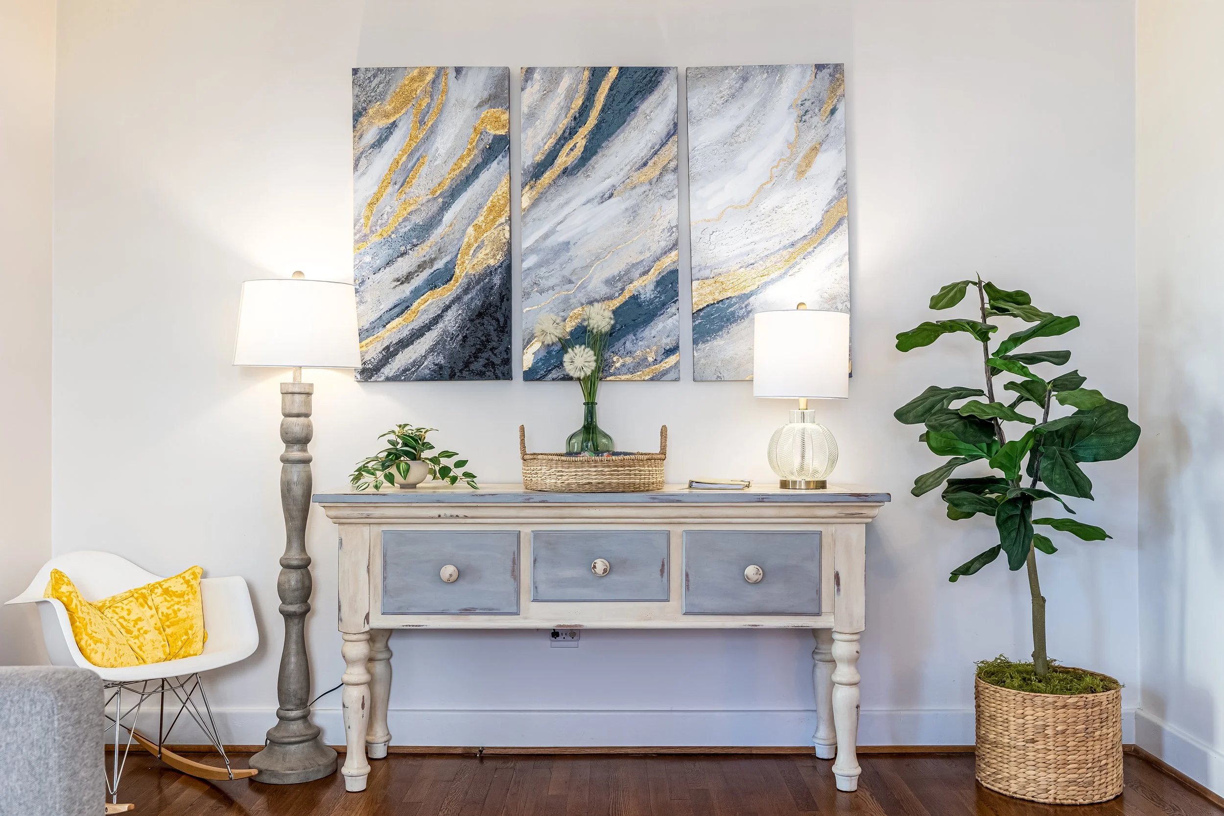 Living room interior with a vintage style sideboard decorated with a glass vase of dandelions and a tray, flanked by two table lamps, with abstract wall art above, a white chair with a yellow cushion, and a tall potted plant to the right.