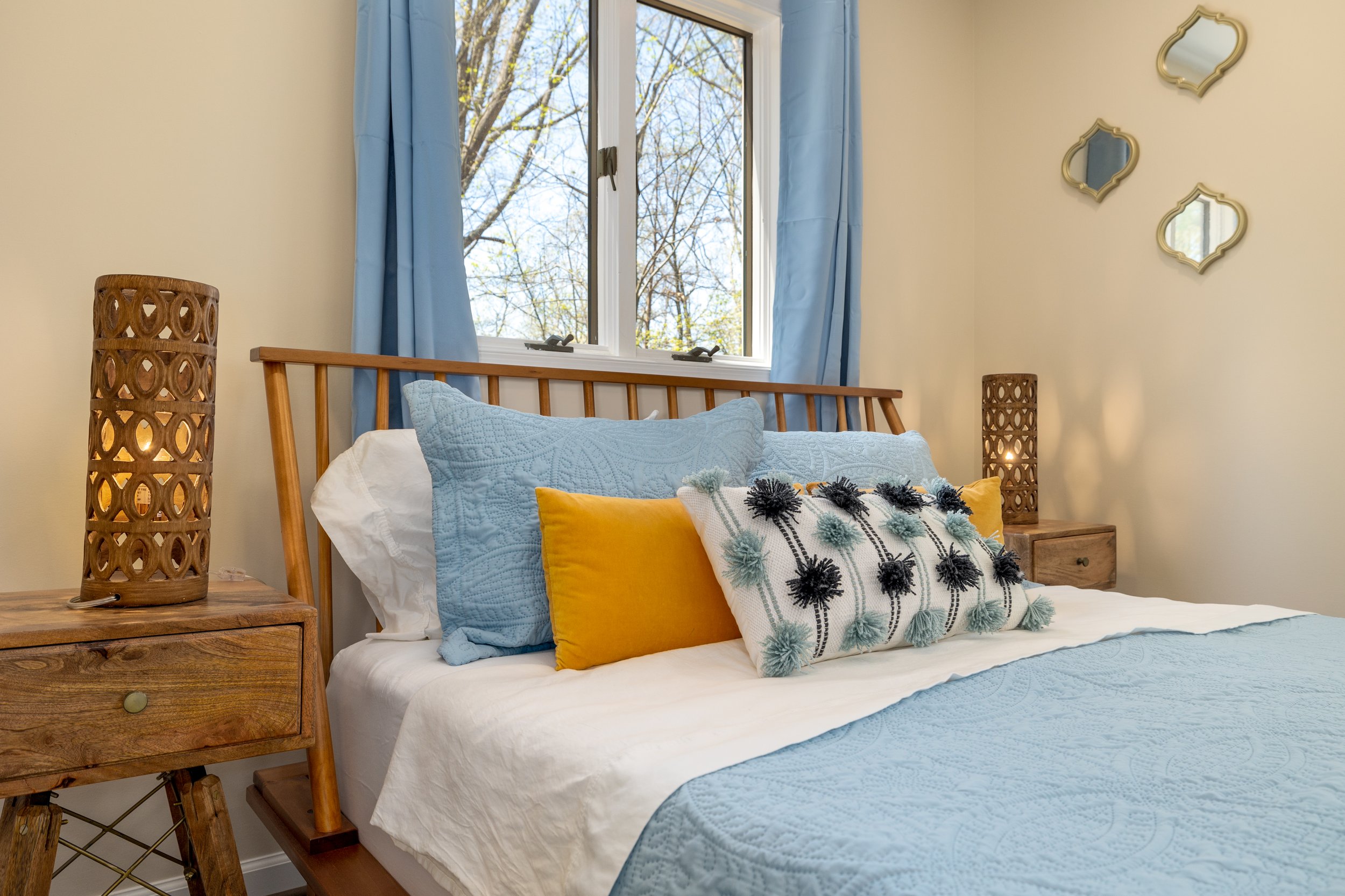 A bedroom with a wooden bed frame, blue and yellow throw pillows, a white quilt, and a window with blue curtains. There are three small mirrors shaped like quatrefoils on the beige wall, a wooden nightstand, and a decorative wooden lamp on the nightstand.