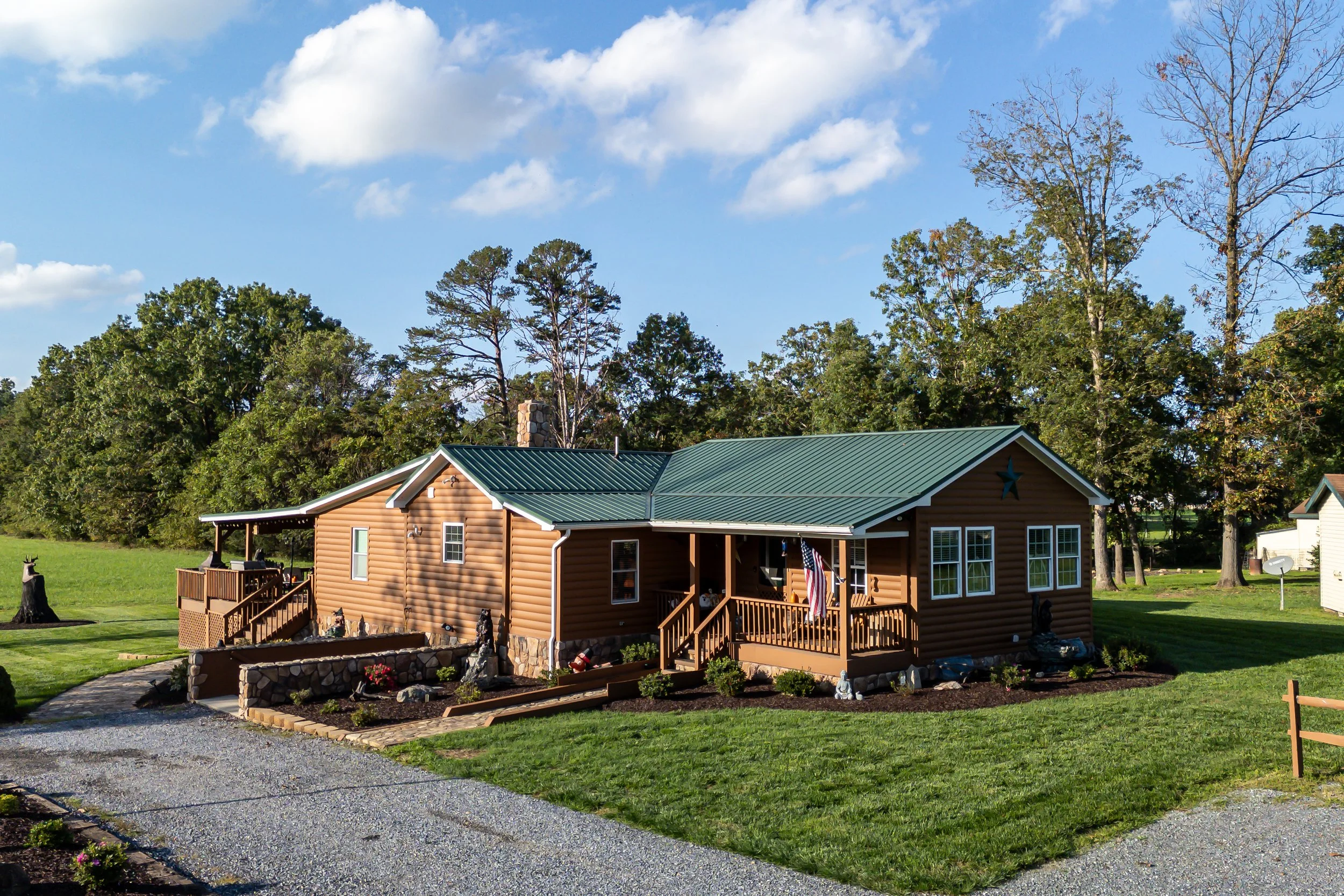 A rustic brown ranch-style house with a green metal roof, surrounded by a well-maintained lawn. The house features a covered porch with an American flag, stone foundation accents, and a gravel driveway. Trees and a detached structure are visible in the background under a blue sky with clouds.