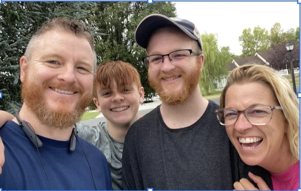 A group of four smiling people, two men and two women, taking a selfie outdoors on a sunny day with trees and houses in the background.