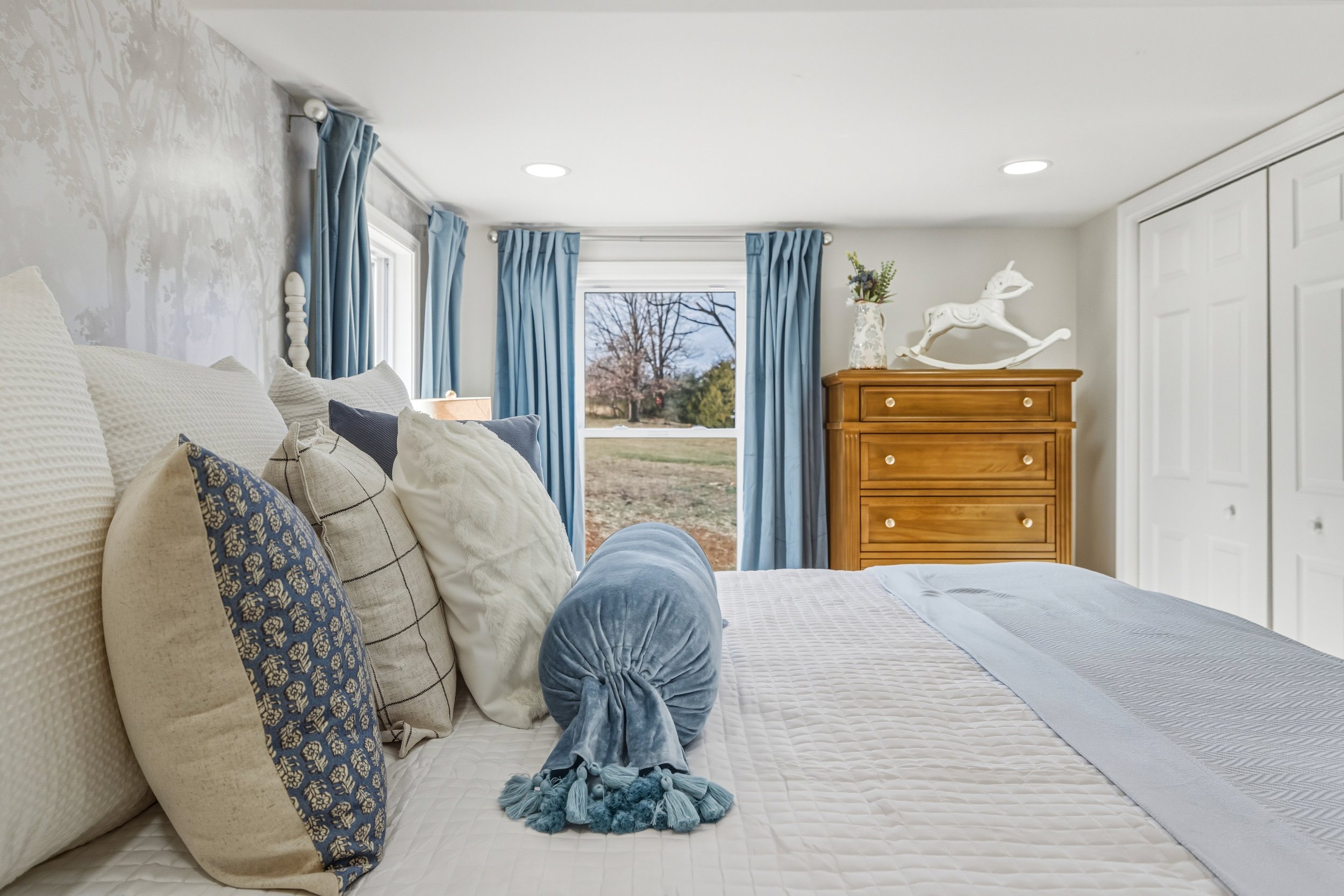 A bedroom with a neatly made bed with multiple pillows, some with patterned and textured covers. There are blue curtains on the windows, tied back with matching fabric. A wooden dresser with a white rocking horse and a floral vase on top is visible, 