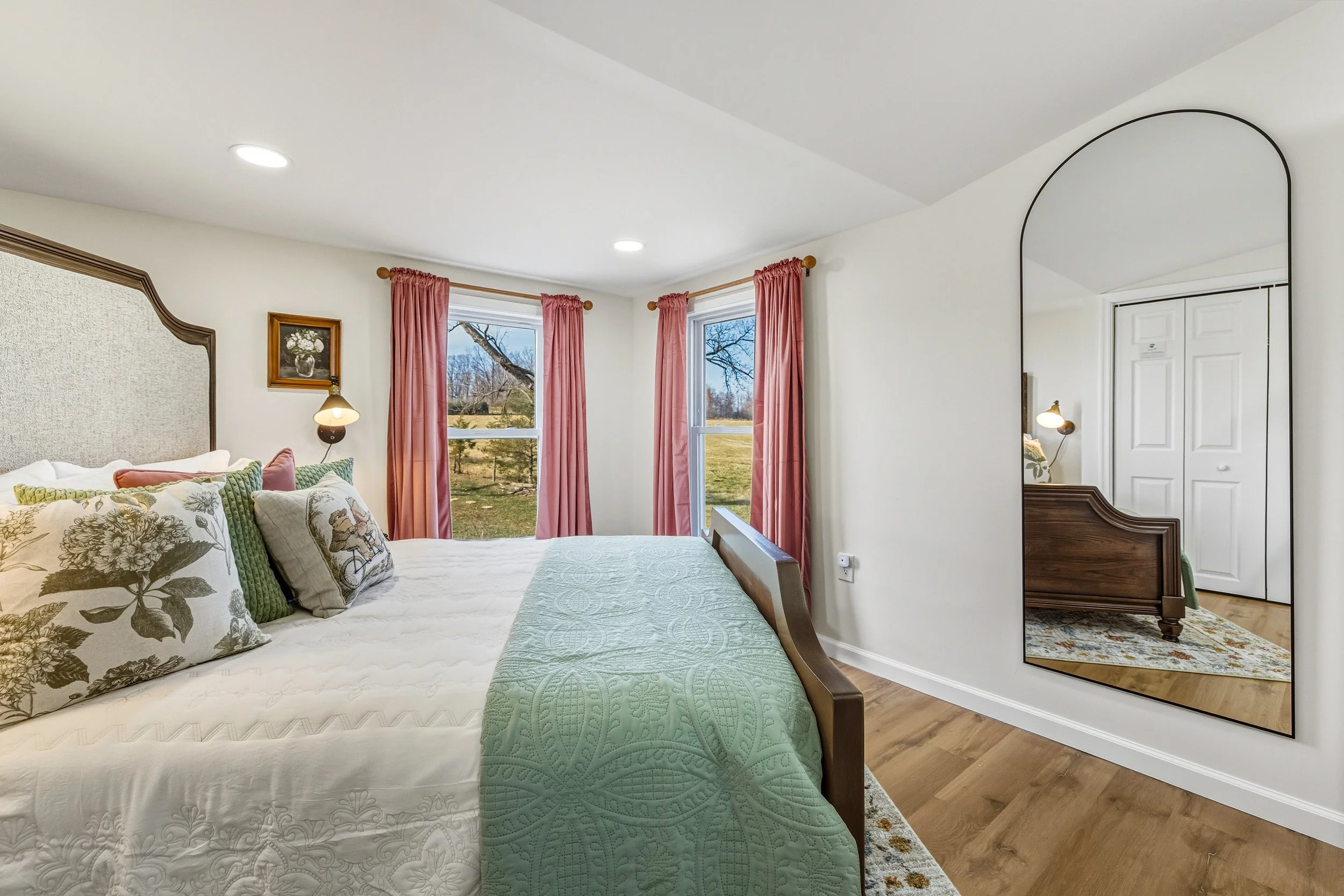 A bedroom with a bed adorned with floral and green pillows, a quilted bed cover, and a large mirror reflecting a white door and a small bench, with windows showing a landscape outside.