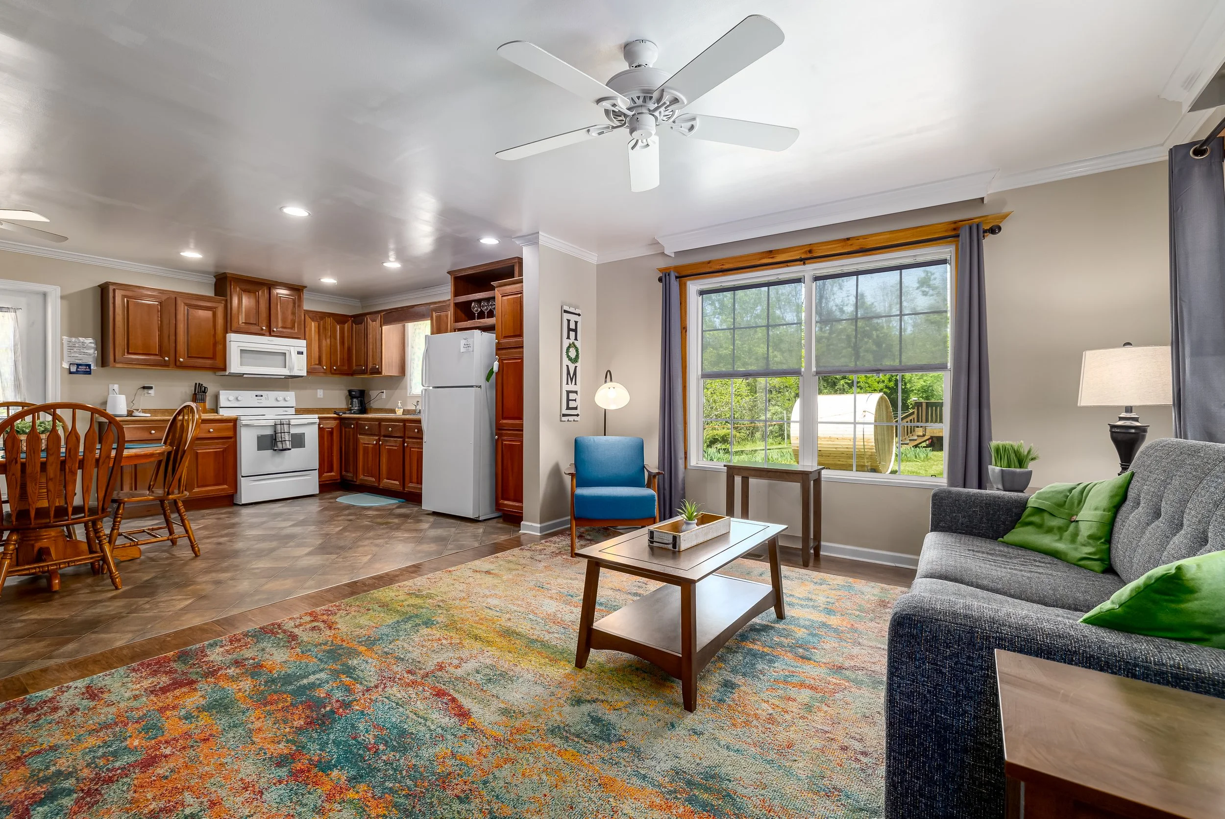 A cozy living room with a large window, gray sofa with green pillows, a coffee table, and a colorful rug. Adjacent to the living area is a kitchen with wooden cabinets, a white stove, microwave, refrigerator, and a dining table with wooden chairs. There is a blue chair and a lamp near the window, and the room has a ceiling fan and recessed lighting.