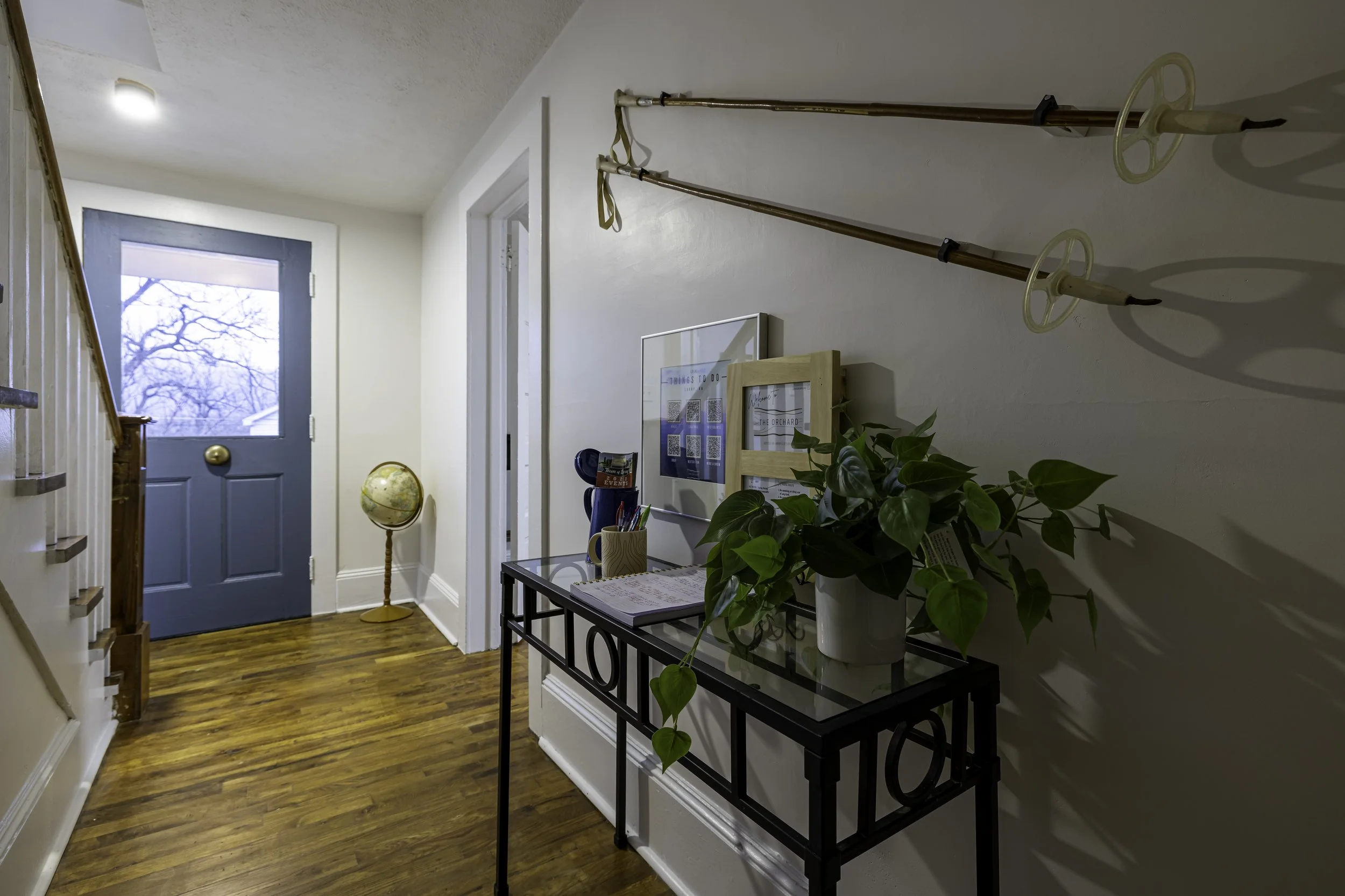 The image depicts a cozy entryway with a blue door, hardwood floors, and white walls. There are two walking poles mounted on the wall, a small table with a potted plant, a globe, and some artwork and framed pictures hanging on the wall. There is a window next to the door letting in natural light.