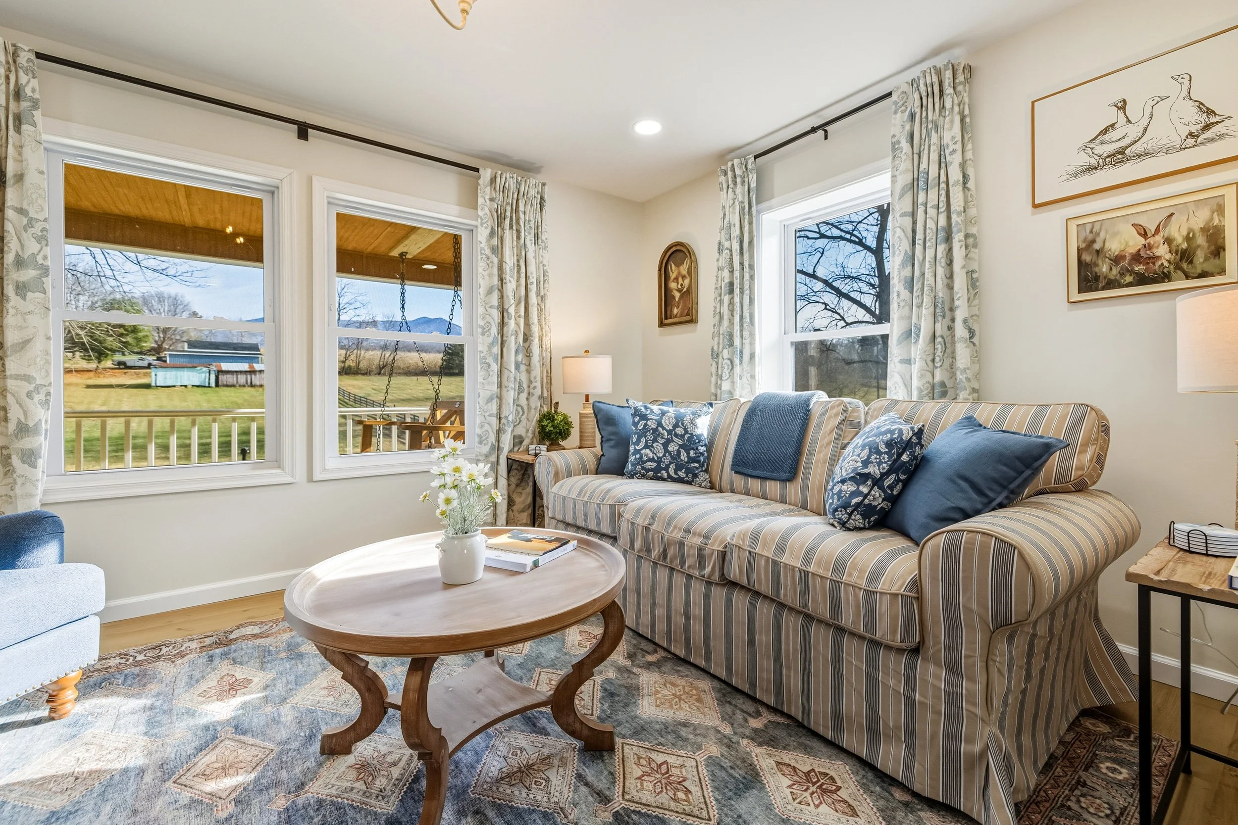 Living room with striped sofa, blue cushions, coffee table with vase of flowers, large windows showing outdoor yard, framed animal artwork on wall.
