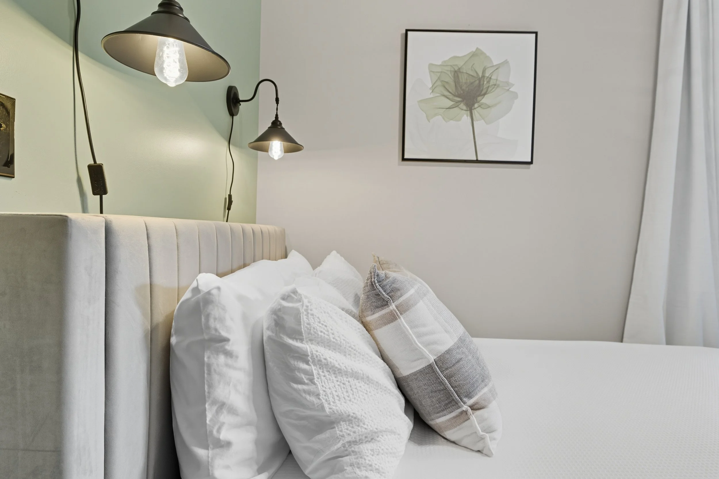 A bedroom scene featuring a bed with white pillows and a plaid pillow, a light green wall with two black reading lamps, a white wall with a framed digital art print of a flower, and sheer white curtains to the right.