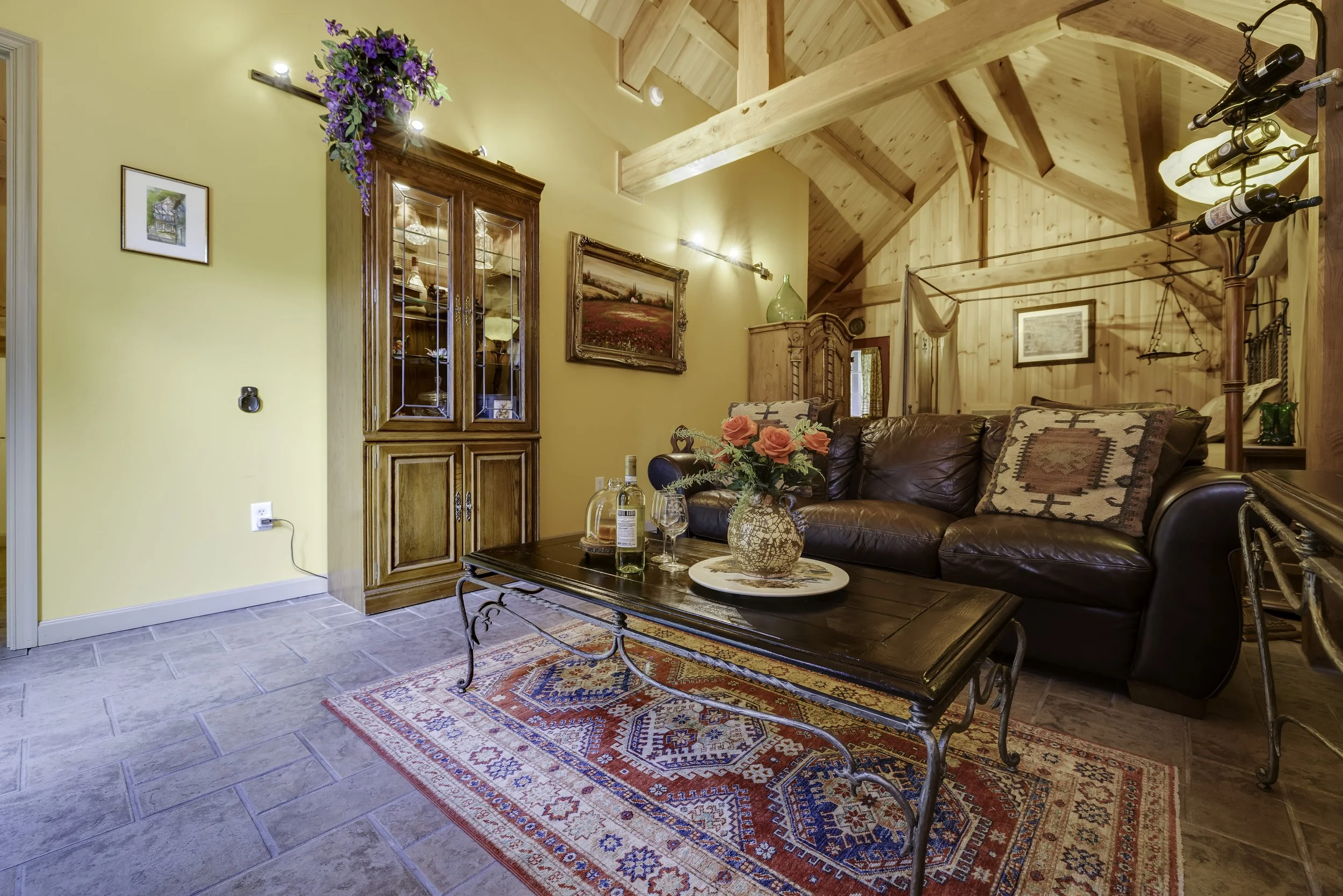 Interior view of a cozy living room with yellow walls, wooden ceiling beams, and a mix of vintage and rustic furniture. Features a leather sofa, a wooden coffee table with a vase of pink flowers, and a colorful area rug. The room has wall art, a wooden cabinet with glass doors, and decorative lighting.