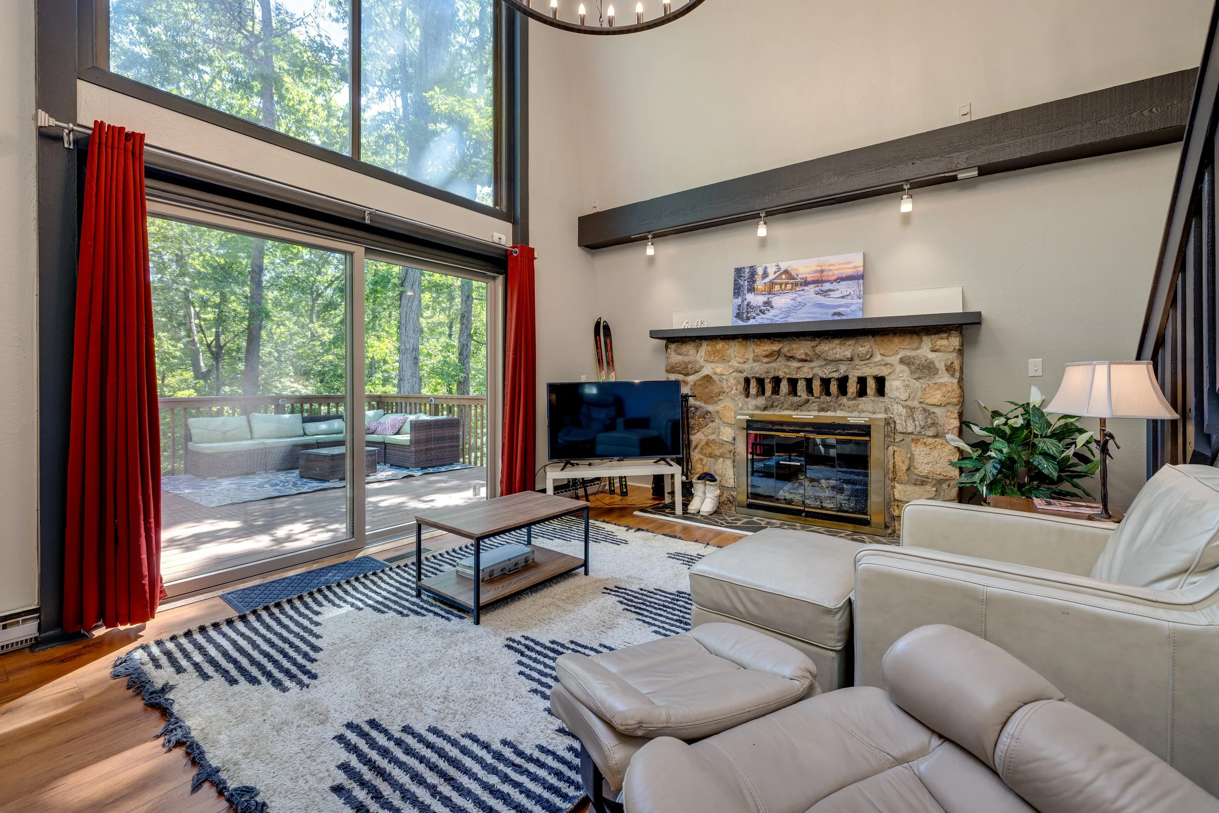 A cozy living room with a white leather sectional sofa and matching ottoman, black and white striped rug, and a stone fireplace decorated with a winter scene painting. Large windows with red curtains reveal a wooded outdoor patio area with outdoor fu