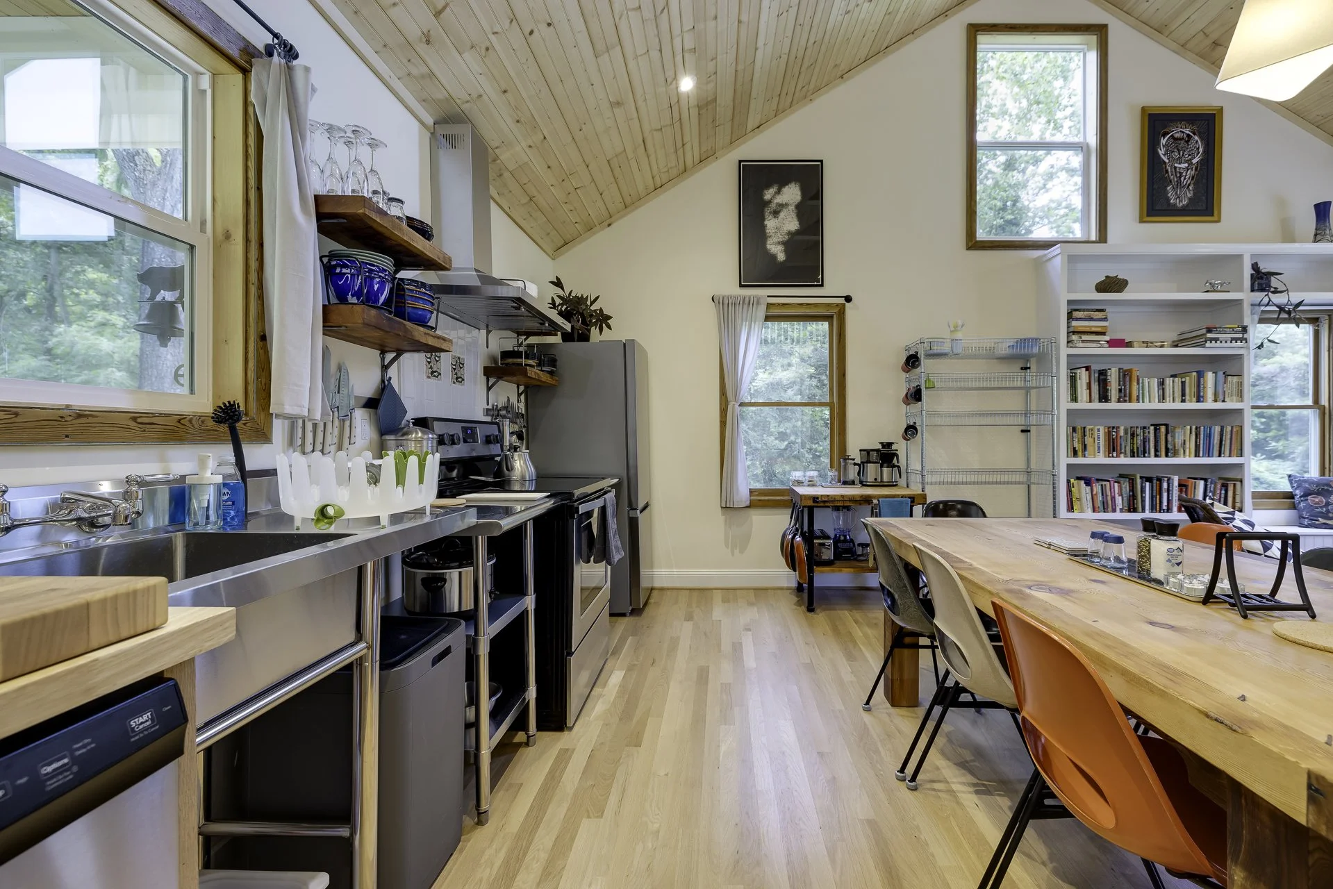 A cozy kitchen and dining area with wooden ceiling and floor, large windows with white curtains, shelves with glassware and dishes, a kitchen stove, and a dining table with multiple colorful chairs.