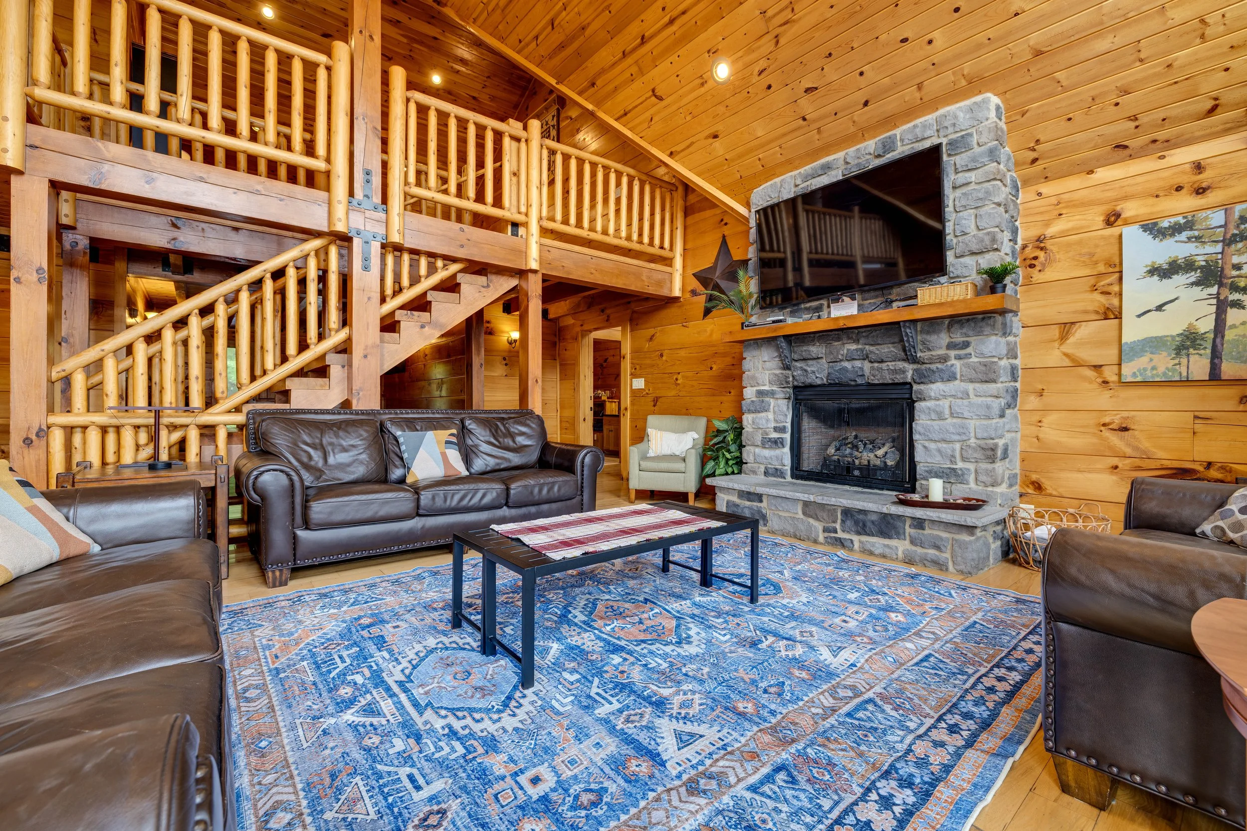 Living room with wooden walls and ceiling, stone fireplace, flat-screen TV, leather sofa, patterned area rug, and decorative items.