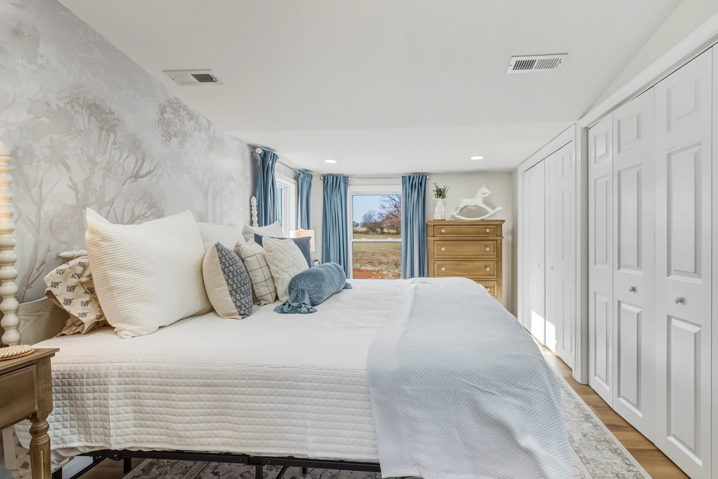A cozy bedroom with a neatly made bed, adorned with white and patterned pillows, and a wooden nightstand. The wall has a subtle tree mural, and there are large windows with blue curtains allowing natural light. A wooden dresser with a decorative hors