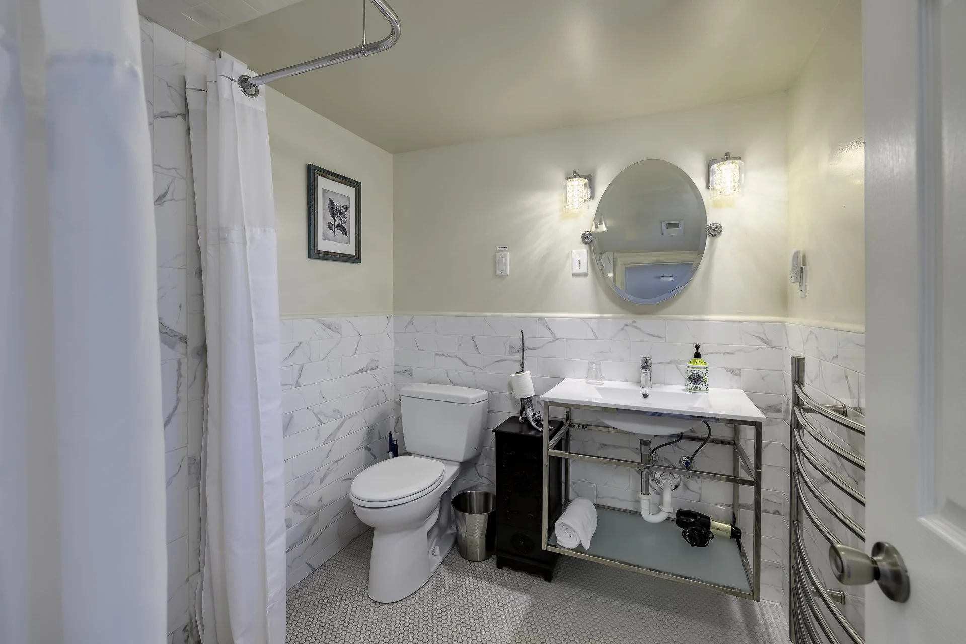 A bathroom with a toilet, a mirror, a sink with a soap dispenser, a small black cabinet, a towel rack, and a framed picture on the wall. The shower area has a white curtain and marble-patterned tiles.