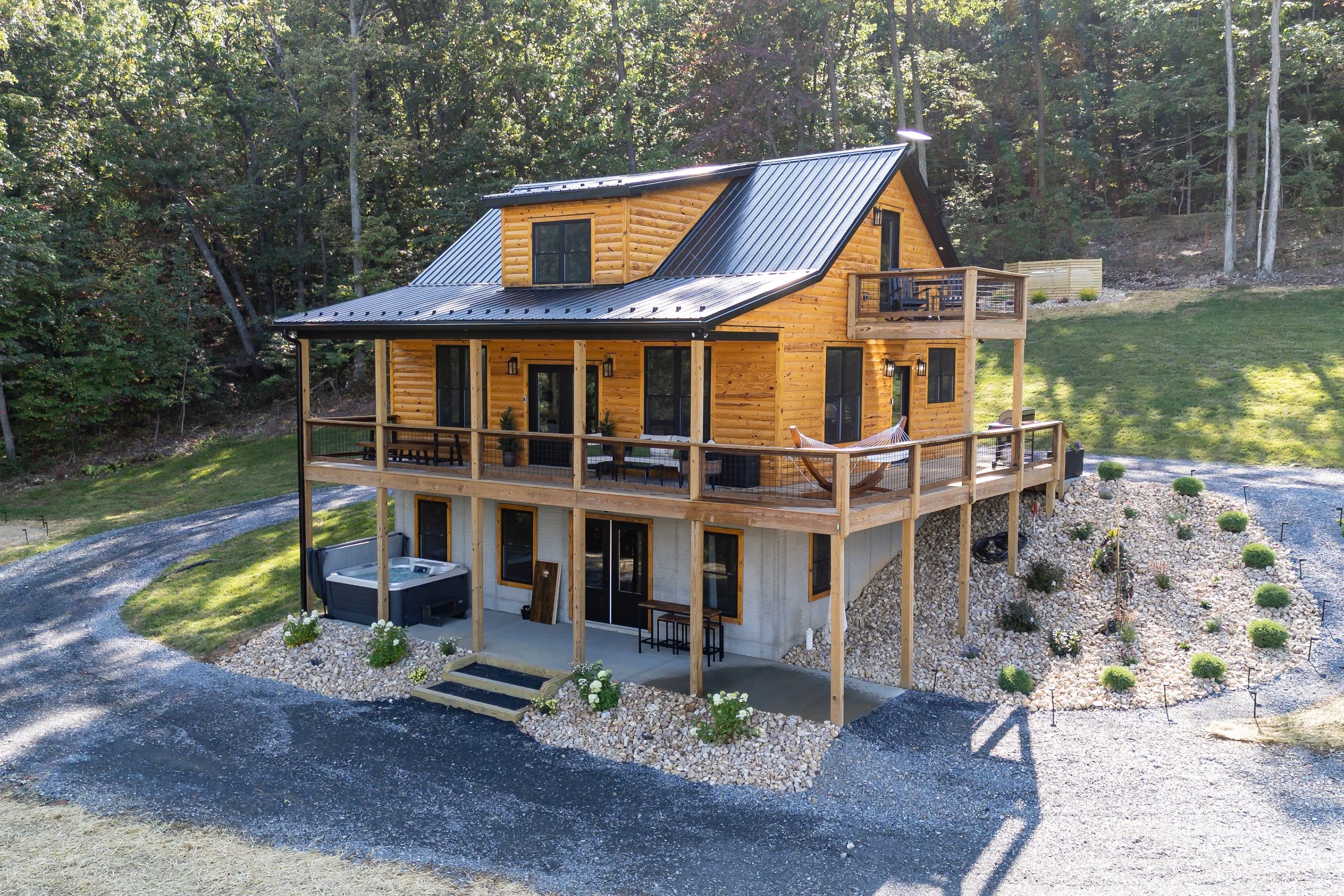 A multi-story wooden house with a modern design featuring a metal roof and large windows, situated in a wooded area with a gravel driveway and landscaped yard.