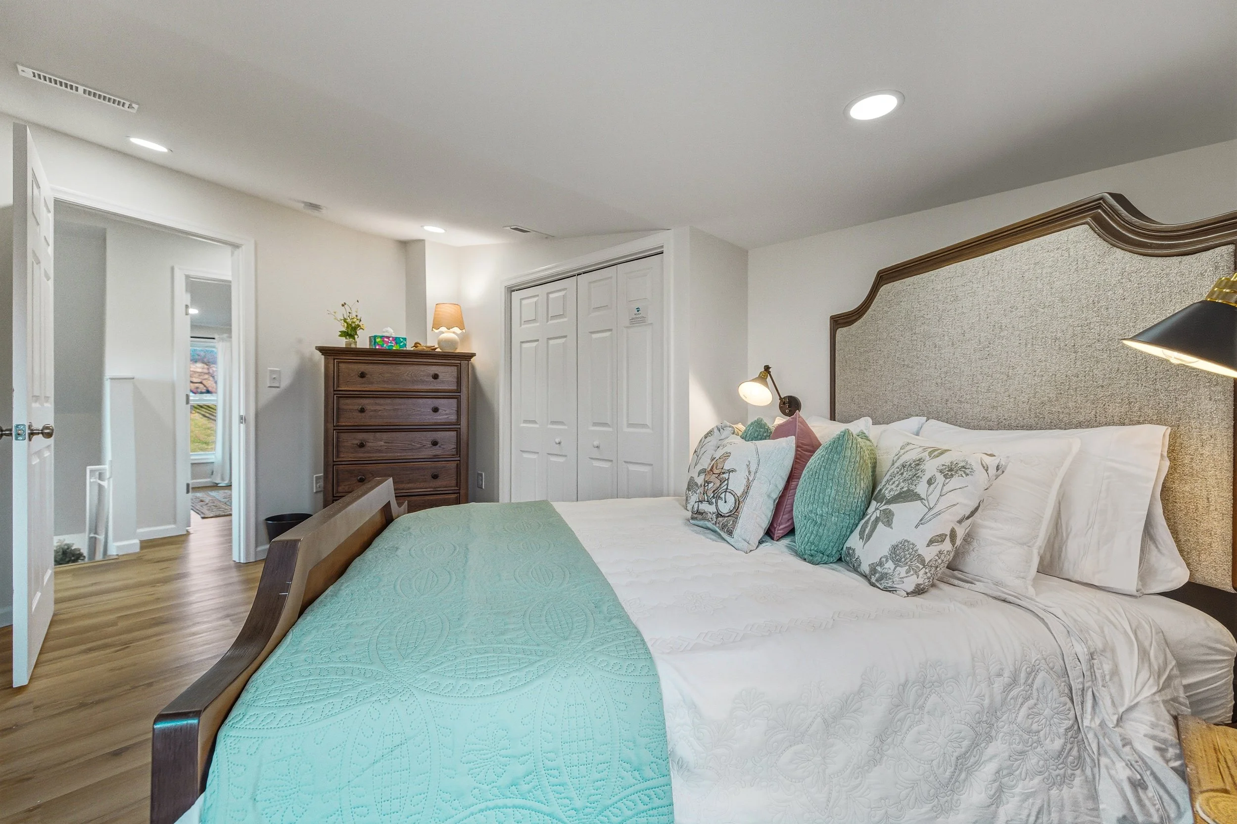 A bedroom with a large bed decorated with multiple pillows, a quilted teal blanket, and a headboard. There are bedside lamps, a dresser with a lamp and small decorations, and a double closet with white doors. The room has wood flooring and white wall