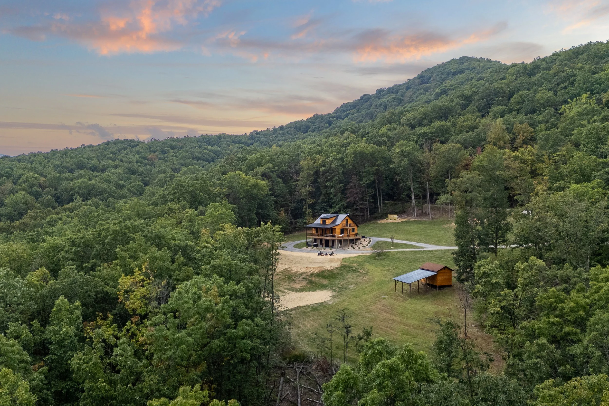 A wooden house on a hill surrounded by trees with a sunset sky in the background.
