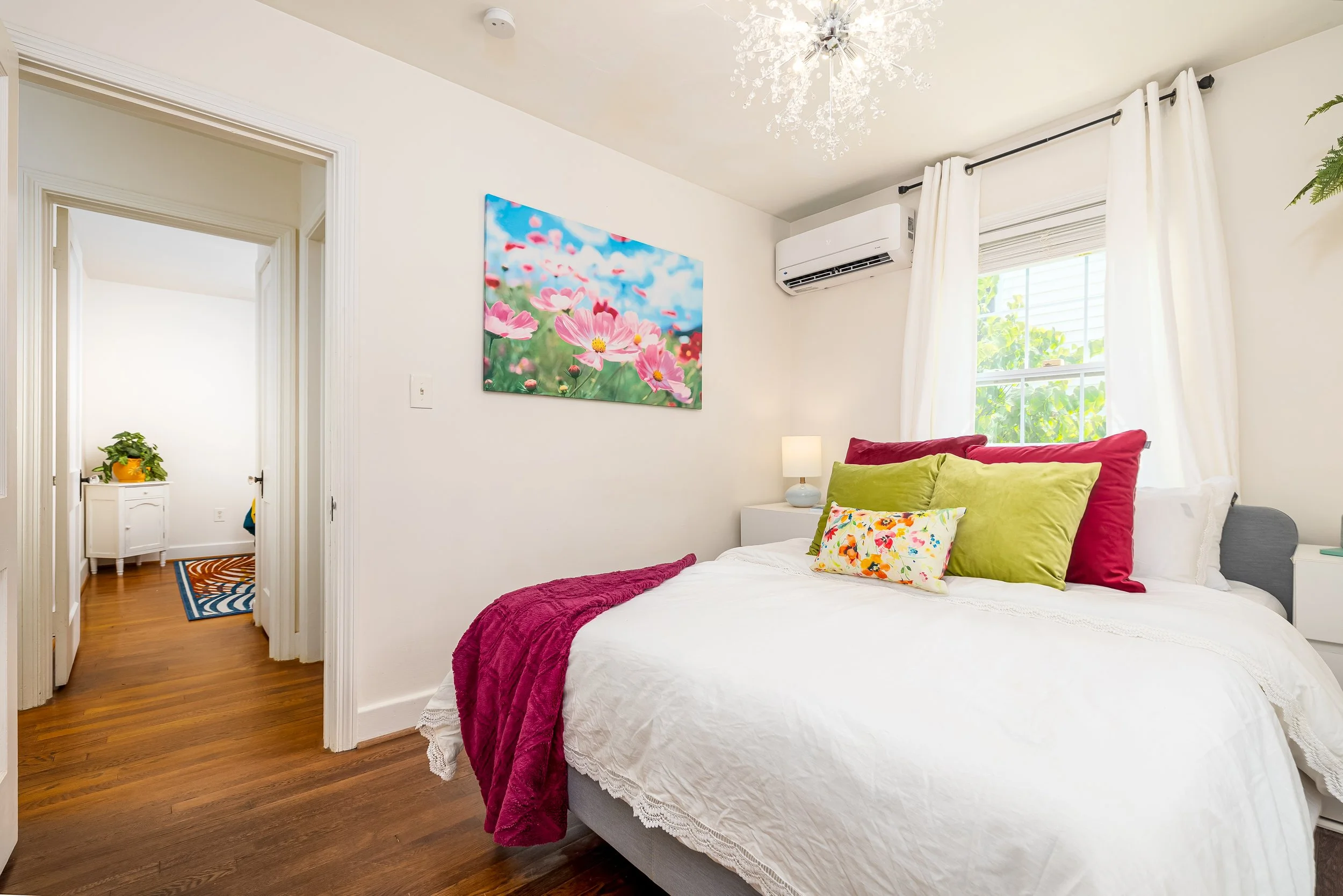 A cozy bedroom with a white bedspread, colorful pillows, and a pink blanket. A floral painting hangs on the wall, and windows with white curtains let in natural light. There is an air conditioning unit and a small nightstand with a lamp. A chandelier