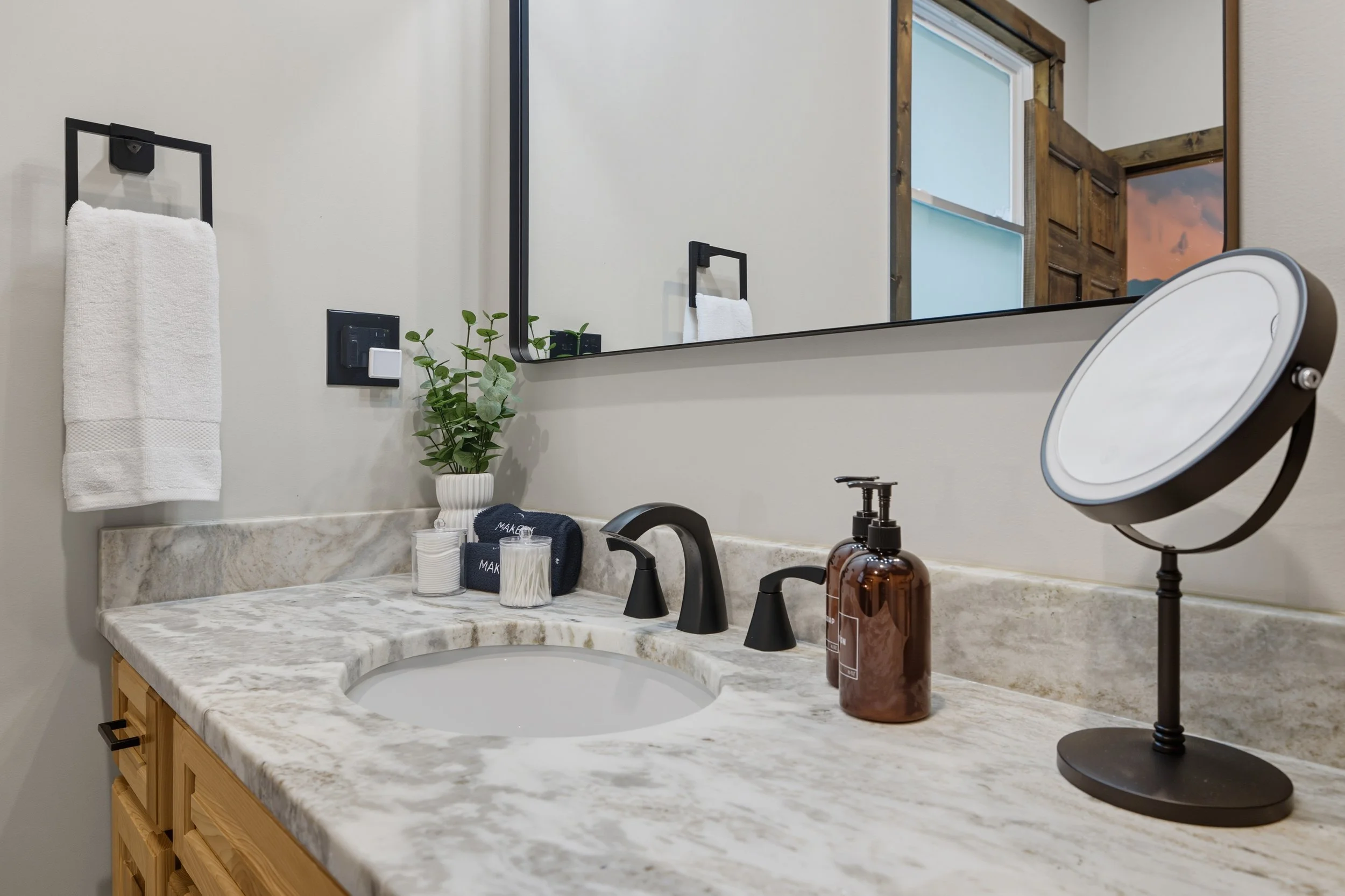 Bathroom vanity with marble countertop, black faucet, mirror, hand towel, green plant, and personal care items.
