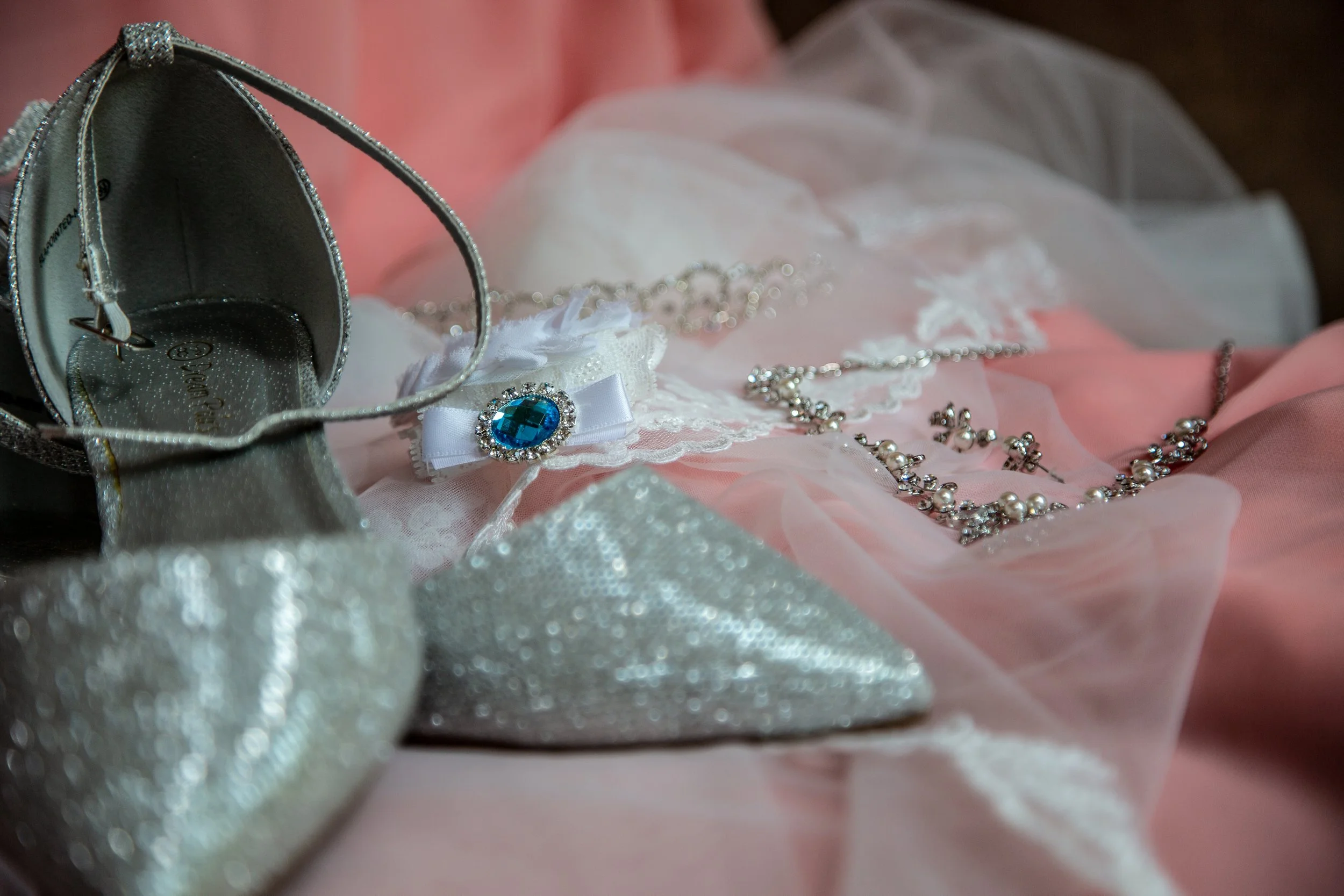 Close-up of a silver and crystal purse, a blue gemstone brooch with rhinestones, pearl necklaces, and pink and white lace fabric accessories arranged on pink fabric.