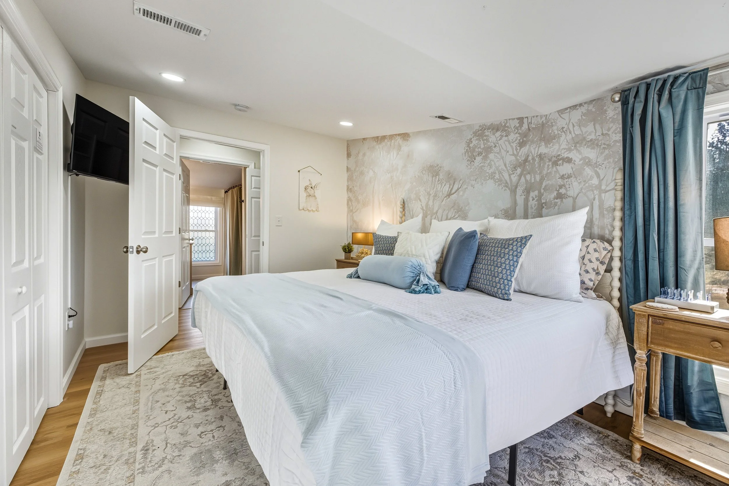 A bedroom with a white bed, blue pillows, a wooden nightstand, a beige wall with tree mural, a window with blue curtains, and a wall-mounted TV.