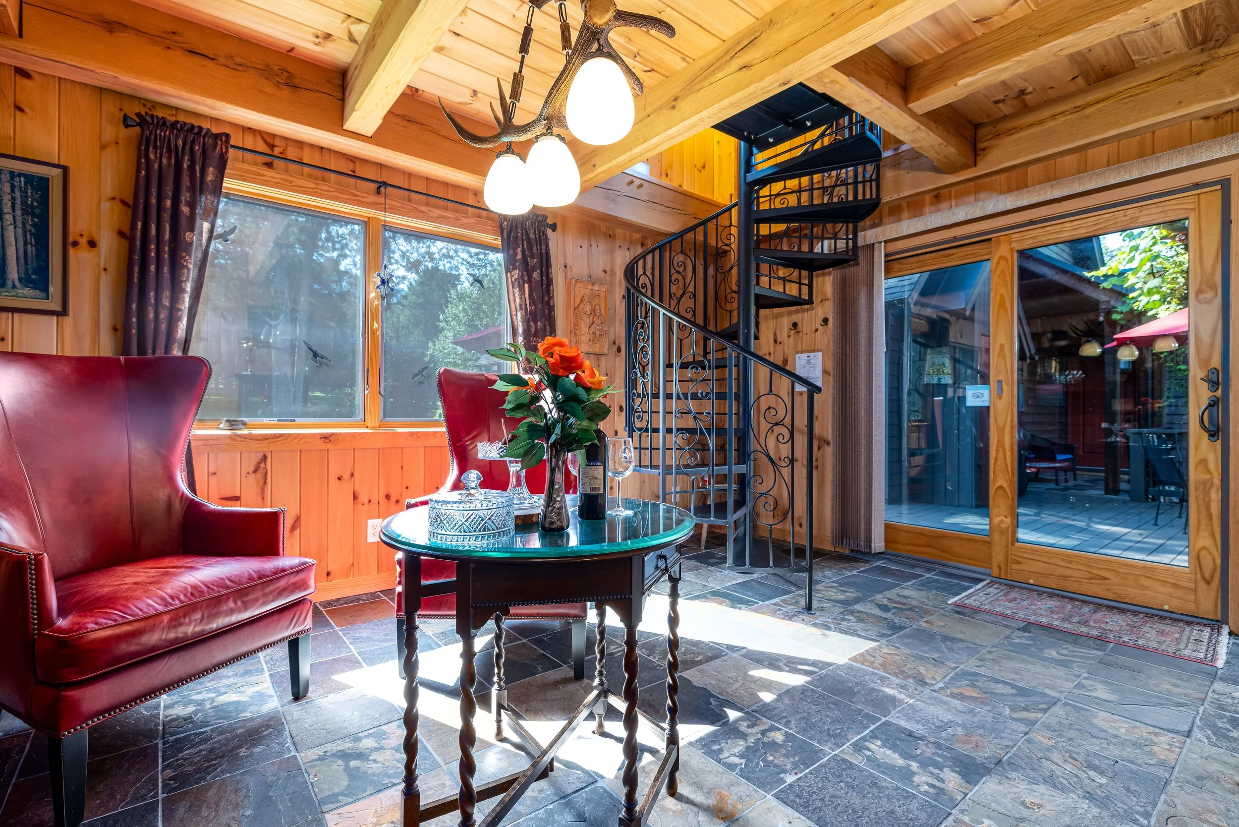 Interior of a wood-paneled living room with a metal spiral staircase, large windows, red leather armchair, glass coffee table with decorative items, flowers, wine bottle, and glassware, and a chandelier with antler design.