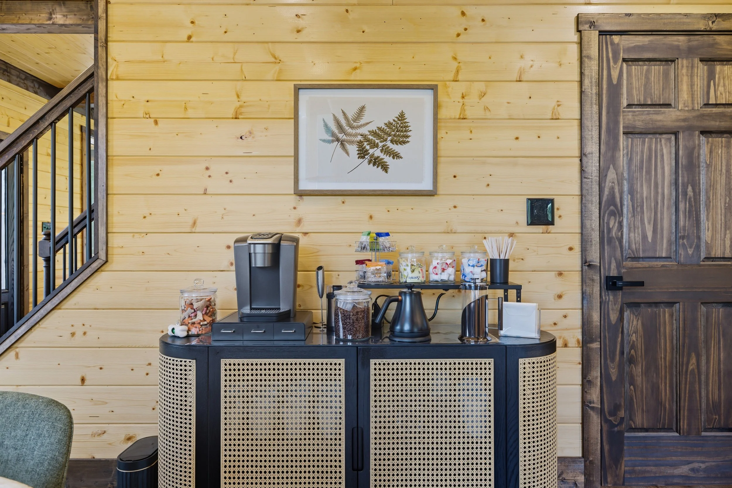 Coffee station with a black cabinet, glass jars of snacks, a coffee machine, and a black kettle, set against a light wooden paneled wall with framed botanical artwork, in a rustic interior.