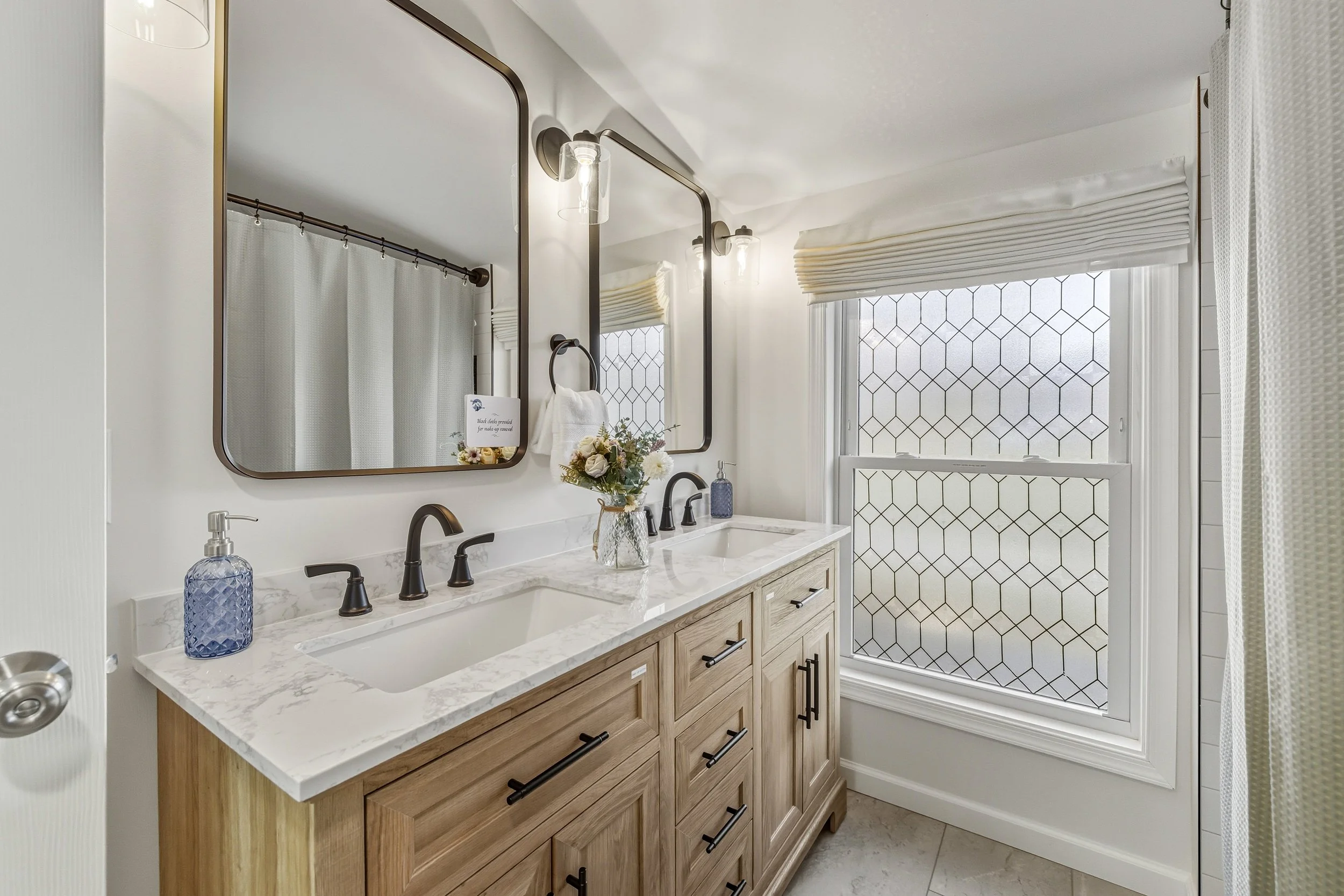A bathroom with a double vanity that has a white marble countertop, black fixtures, and light wood cabinets. Two large mirrors with black frames are mounted above the sinks. There is a decorative flower arrangement on the counter and a soap dispenser
