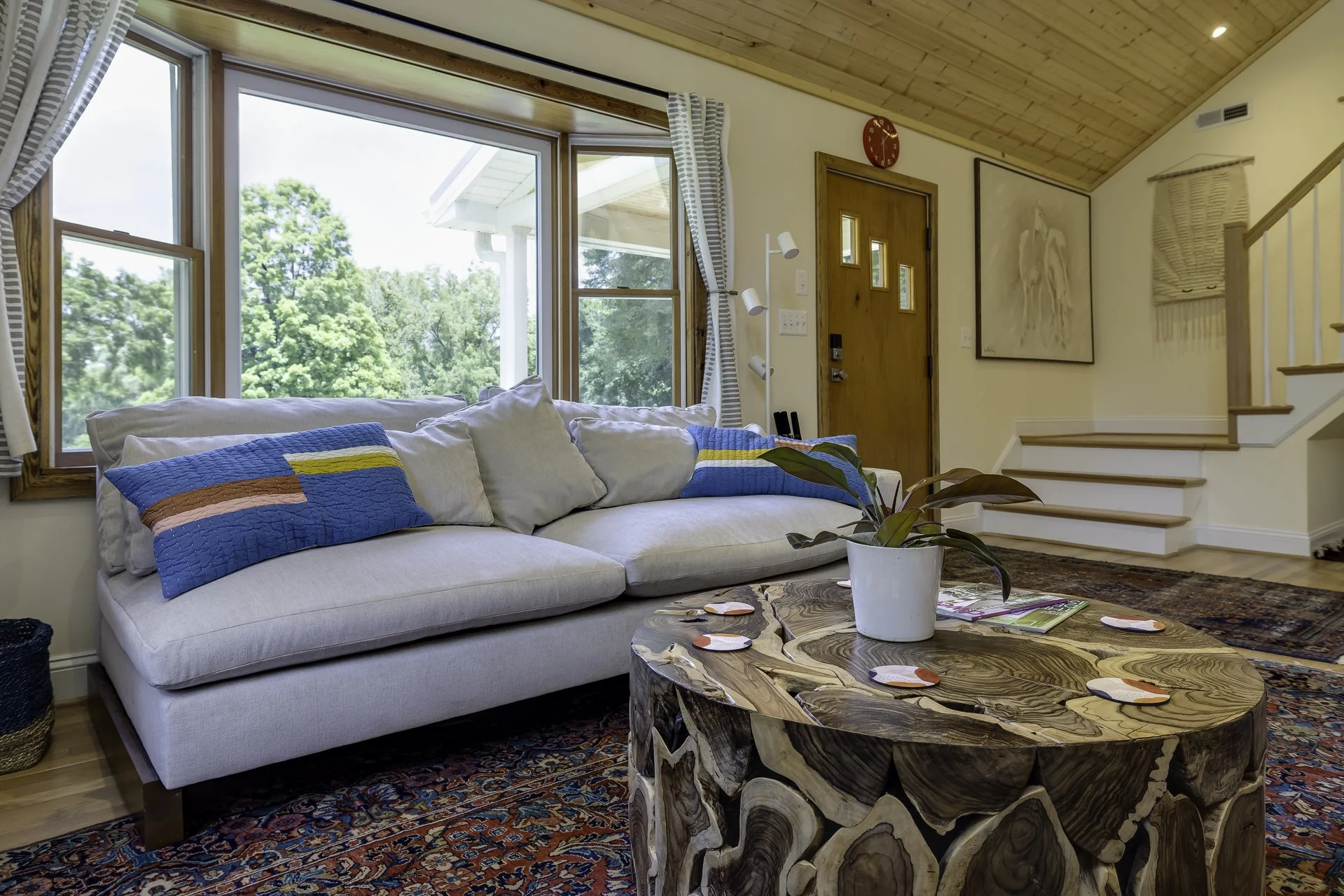 Living room with beige sofa, colorful pillows, wooden coffee table, large windows showing green trees, and staircase.