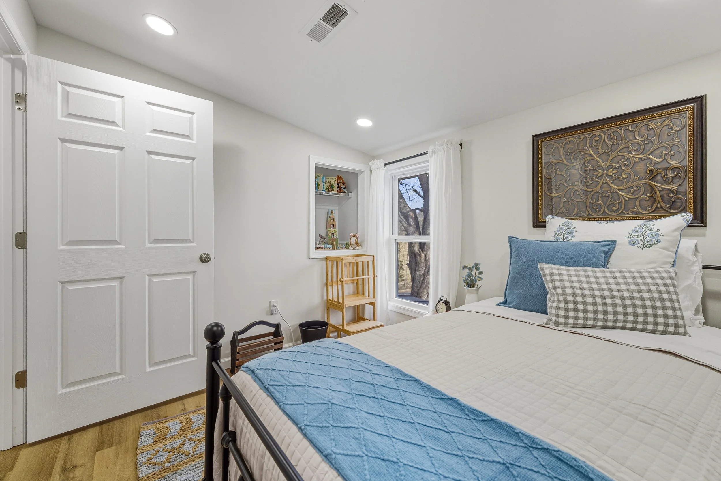 A bedroom with white walls, a bed with pillows, a window with white curtains, a wooden shelf with toys, and a decorative wall panel