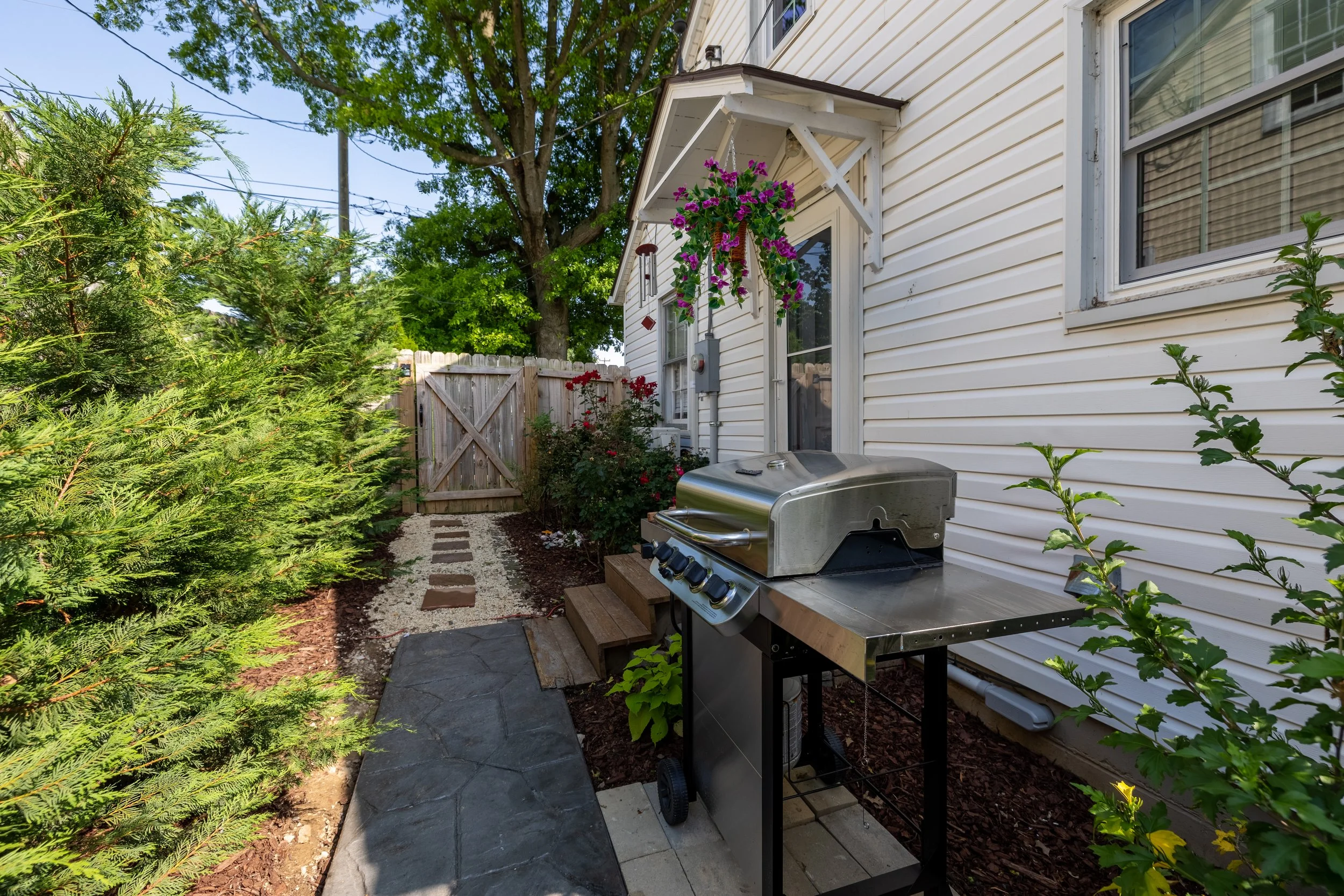 Backyard with small stone pathway, lush green bushes and trees, a white house siding with a window, hanging purple flowers, a stainless steel grill, and a wooden fence.