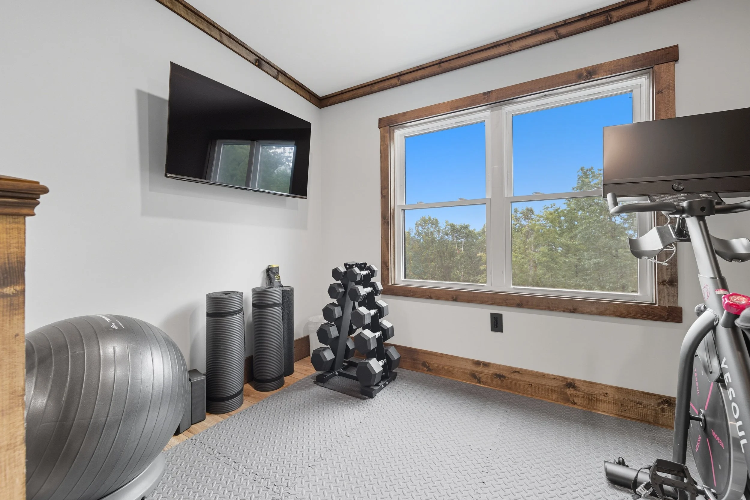 A small home gym with a large exercise ball, foam rollers, dumbbells on a rack, and a stationary bike near a window showing trees and blue sky.