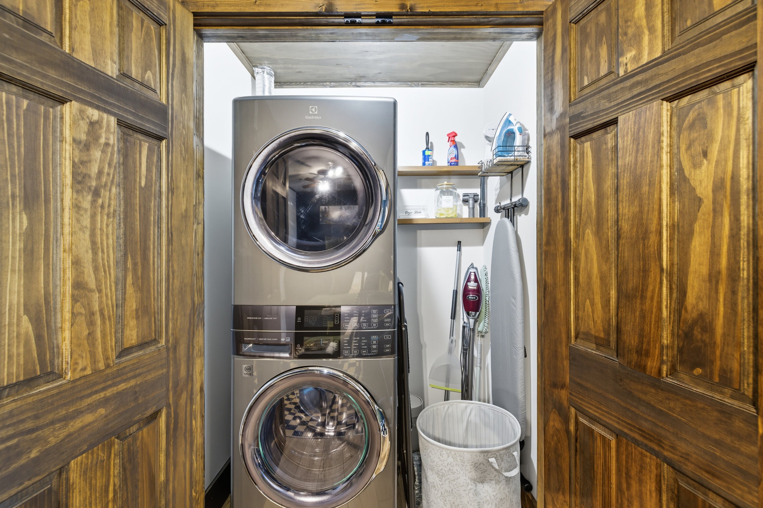 A compact laundry room with a stacked washer and dryer in a small, enclosed space. The washer and dryer are modern, stainless steel Electrolux models. Surrounding the appliances are wooden cabinets on either side, with open shelves above holding clea