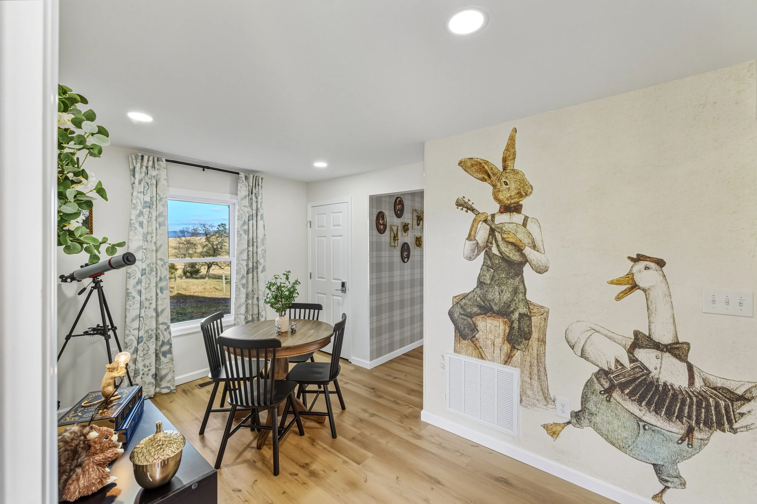Interior of a dining area with a round wooden table, black chairs, a window with floral curtains, and mural art of anthropomorphic animals playing musical instruments on the wall.