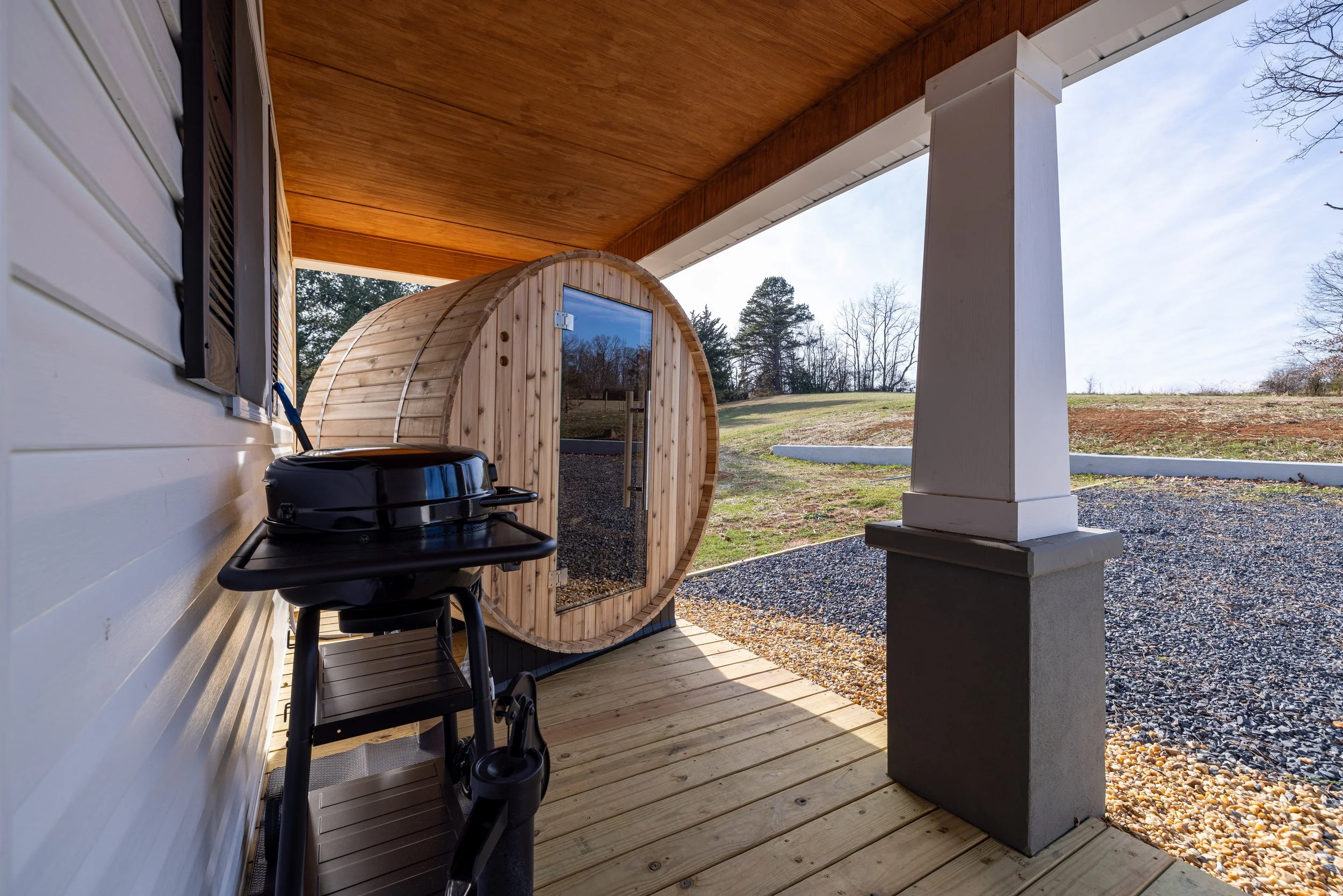 A small wooden sauna on a porch with a black grill and a black chair, overlooking a grassy yard with trees in the distance.