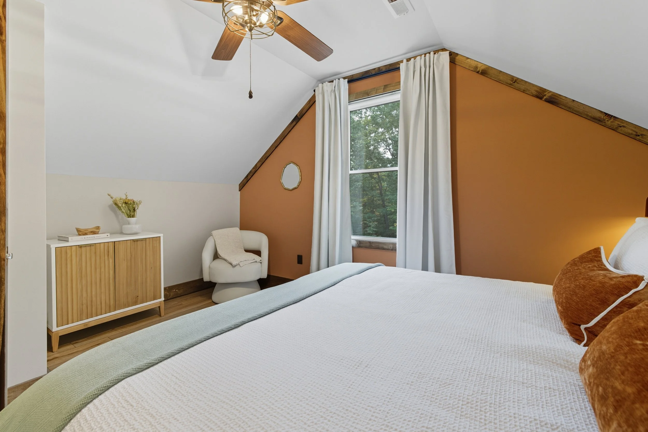 Bedroom with white bedspread, orange accent wall, large window with white curtains, ceiling fan, and light fixture, minimal furniture, and natural lighting.