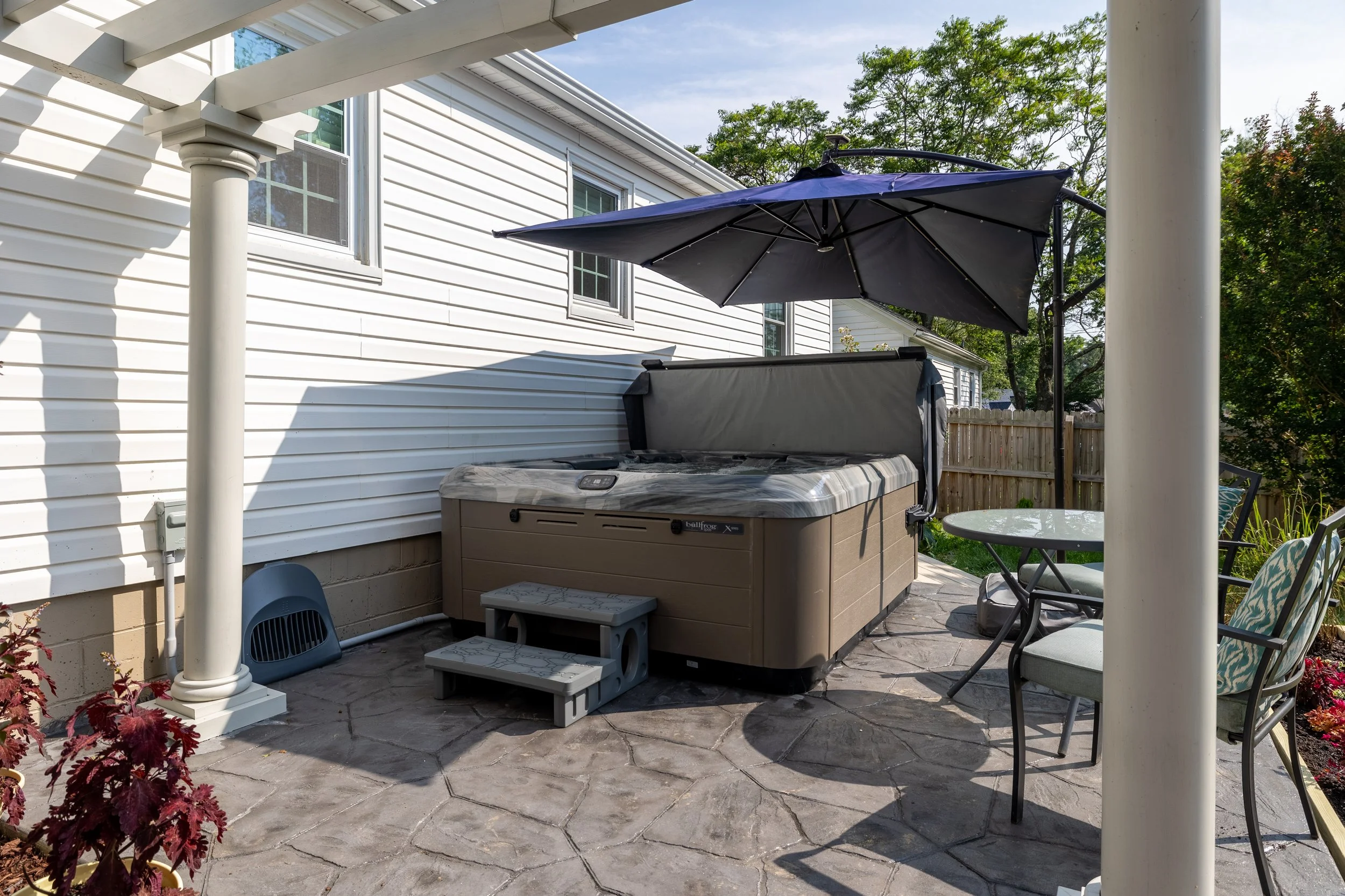 A backyard patio with a hot tub covered by a gray cover, a black patio table with a patterned chair, a blue umbrella, and a white house with windows and trees in the background.