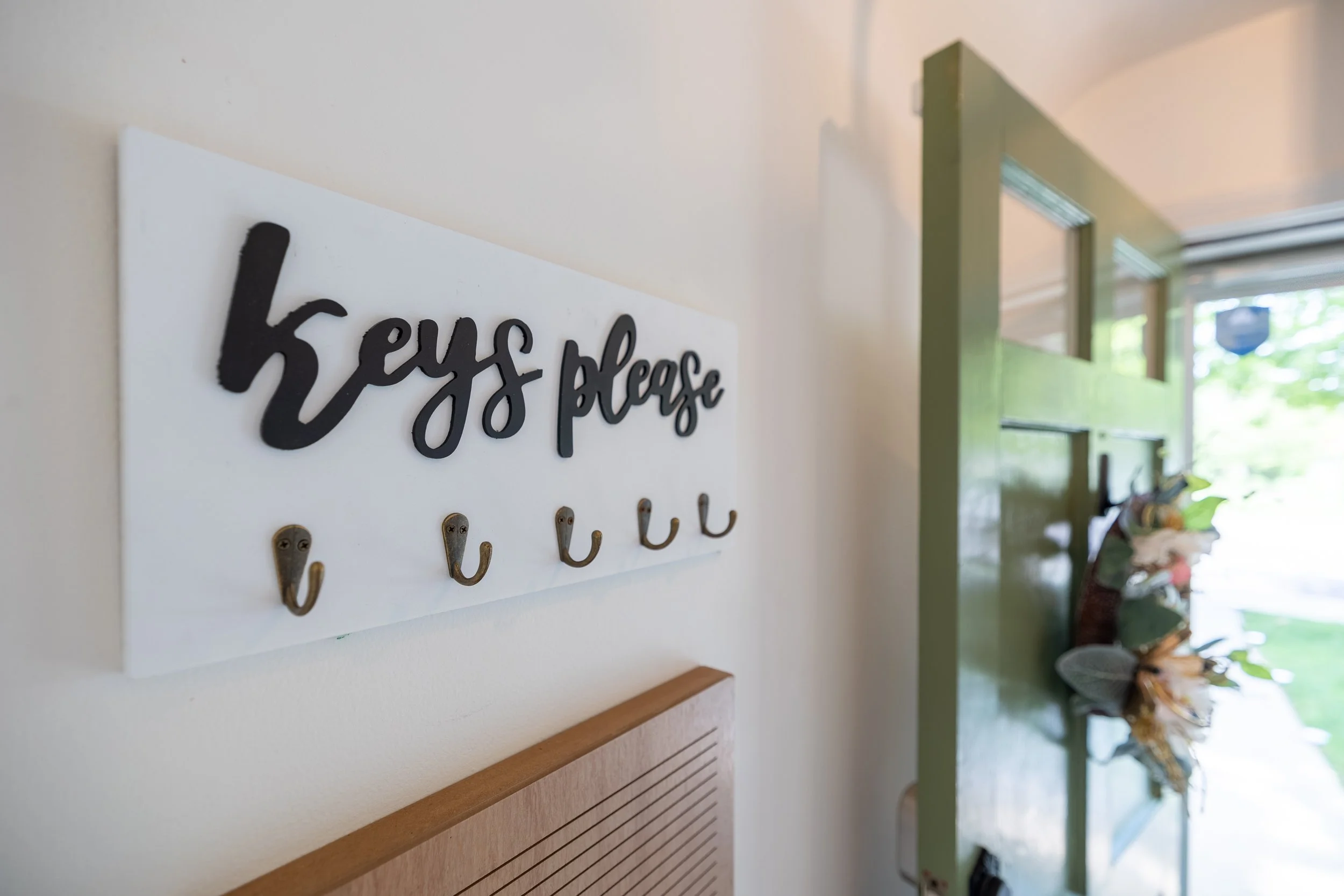 A white wall with black cursive text that reads 'Let's please' and hooks underneath. In the background, there are green walls, windows, and trees outside.