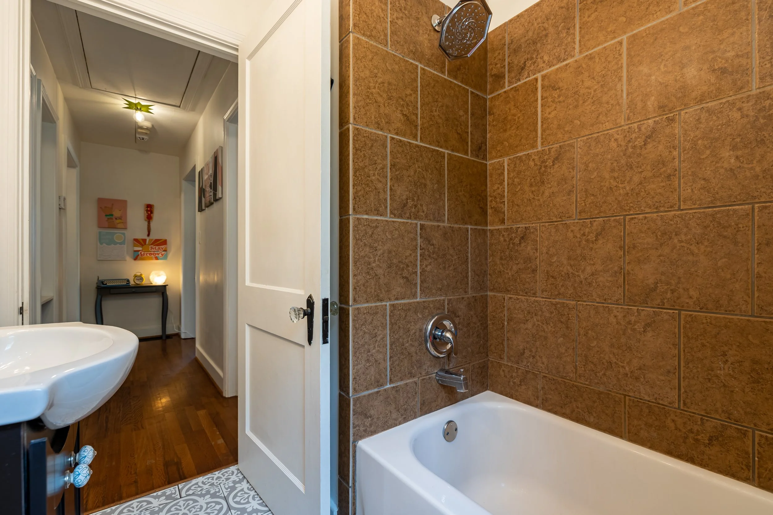 Bathroom with a bathtub, brown tiled wall, and a partial view of a hallway with artwork on the wall.