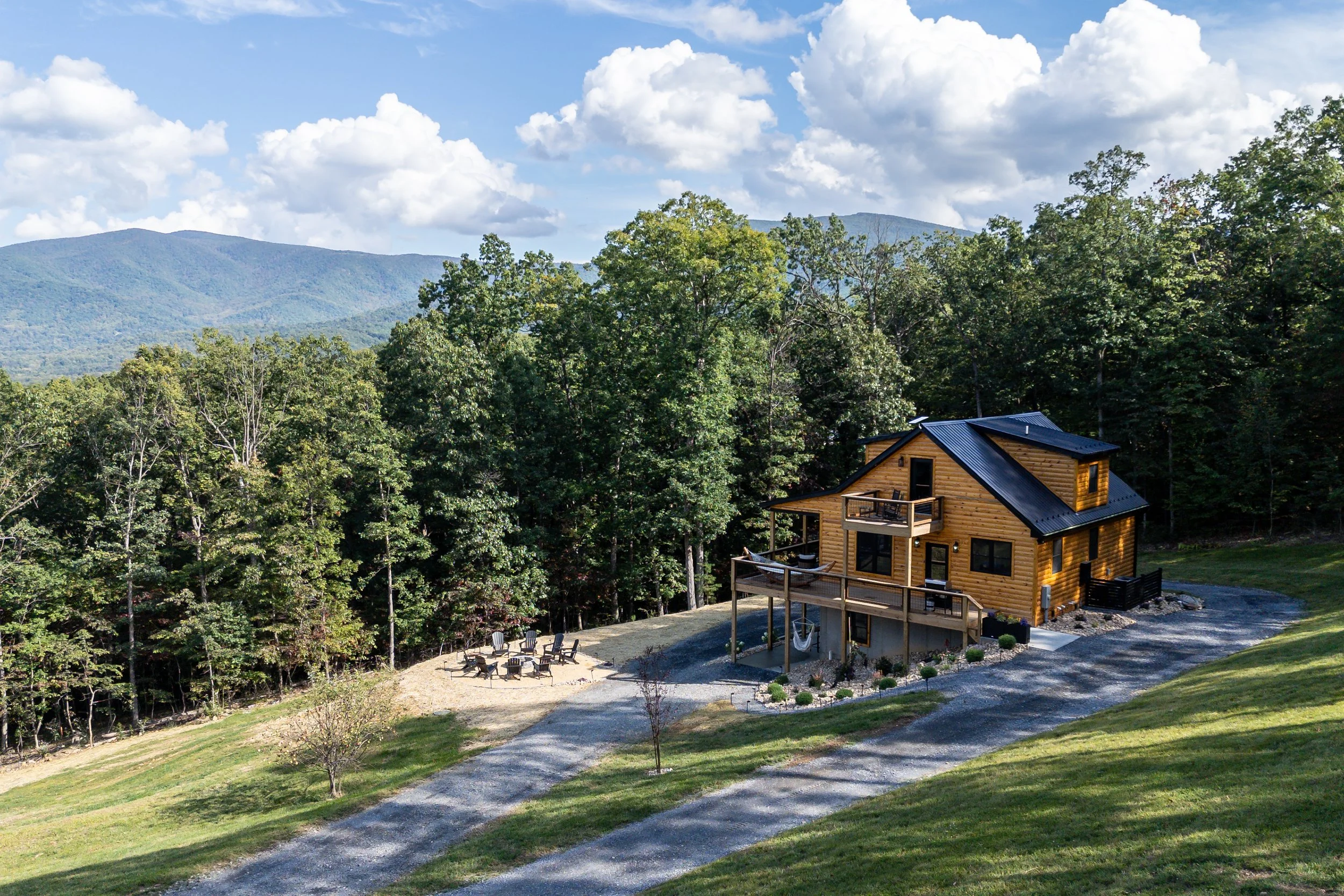 A wooden house with a black roof is set in a lush green landscape with a backdrop of mountains. The house has a deck with outdoor furniture and is surrounded by a gravel driveway and well-kept lawn. The sky is partly cloudy with patches of blue.