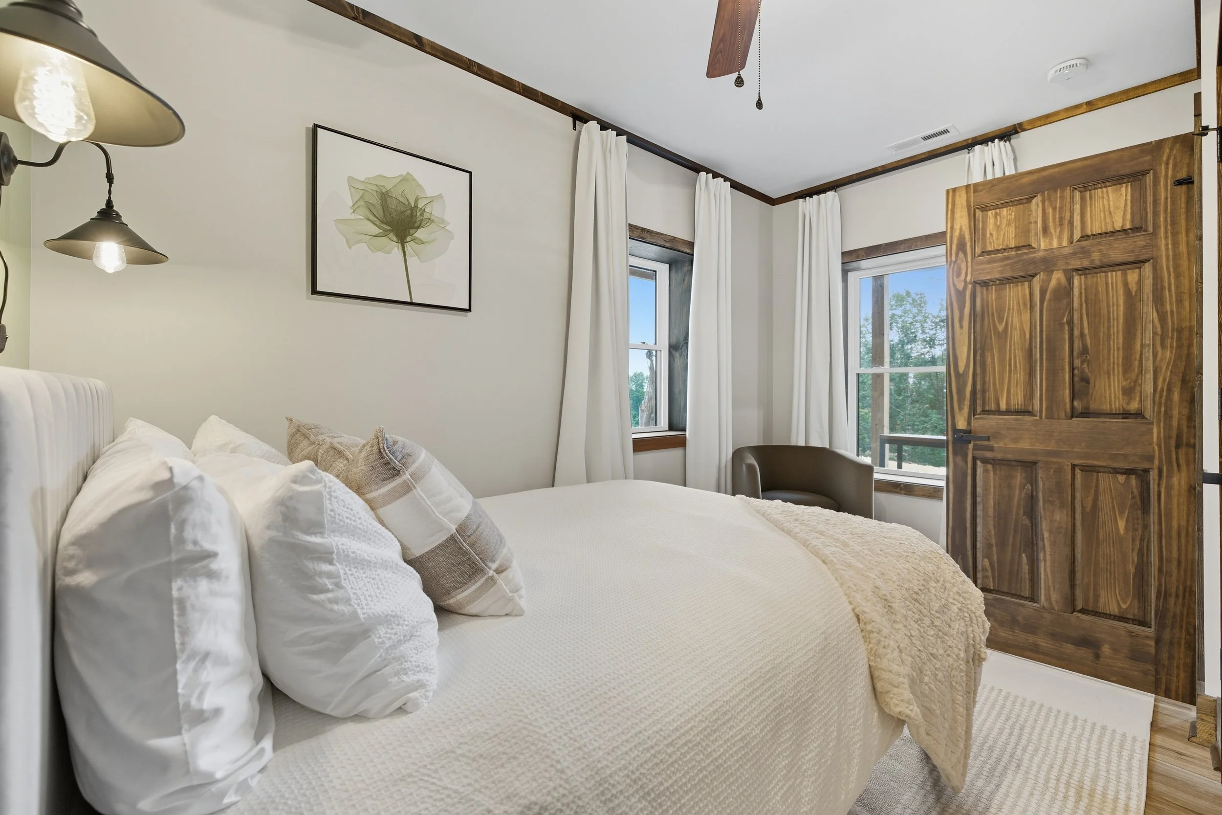 A bedroom with a white bed, beige pillows, and a beige blanket, wooden framed windows with white curtains, a wooden door, a framed flower art piece, a wall sconce lamp, and wooden trim on the ceiling.