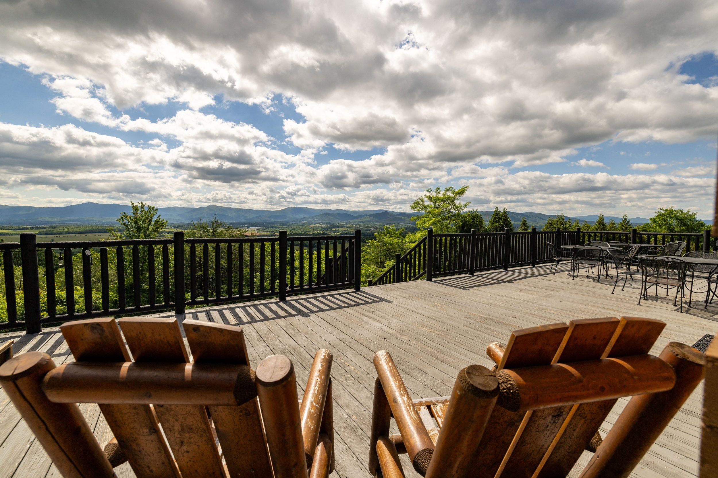 A spacious outdoor wooden deck with outdoor furniture, overlooking a scenic view of green trees, rolling hills, and a partly cloudy sky.
