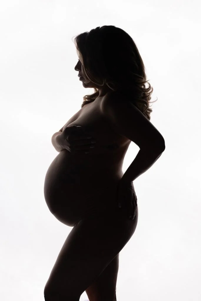 Silhouette of a pregnant woman with wavy hair, standing sideways against a white background, holding her breast with her right hand and resting her left hand on her hip.