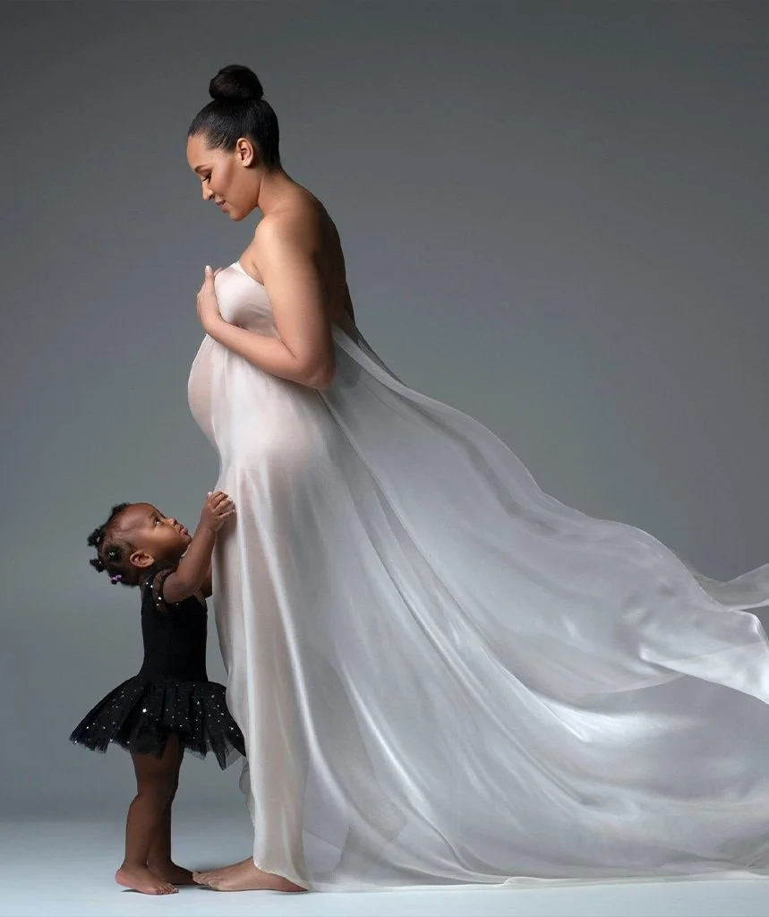 A pregnant mother in a flowing white silk looking down at her young daughter who is reaching up to her. 