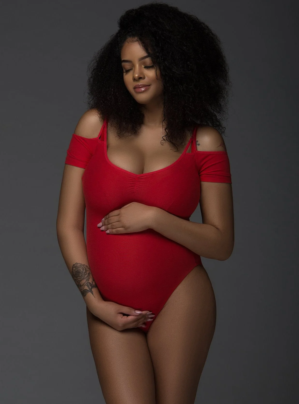 Chicago maternity photography of woman with curly dark hair in a red dress with cold shoulder straps, standing against a dark gray background. She has her eyes closed and her hands are covering her pregnant belly.