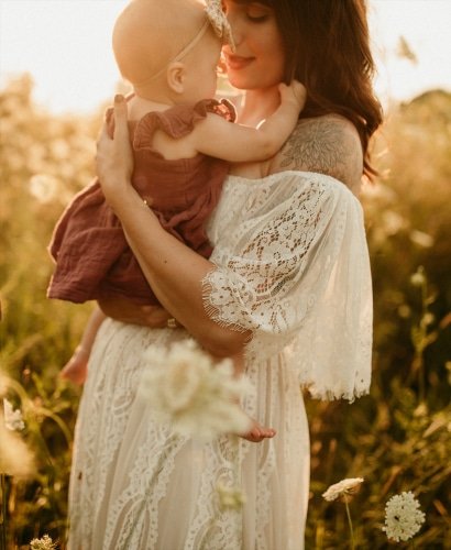 A woman in a white lace dress holds her baby in a field of flowers—captured by a Chicago maternity photographer at golden hour.