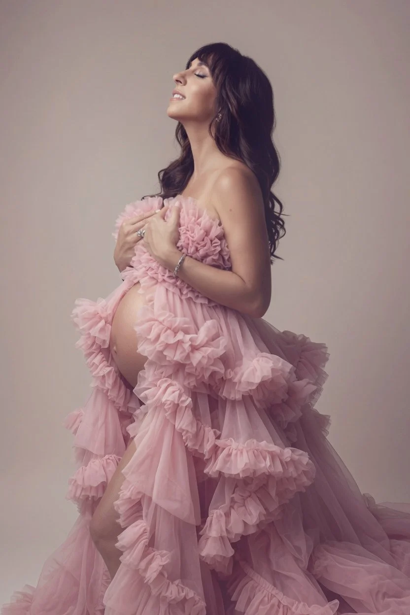A pregnant woman with long dark hair in a pink ruffled dress, holding her chest with a content expression, standing against a plain background.